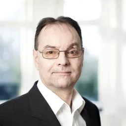 Porträt eines Mannes mit Brille in formellem Anzug vor unscharfem Hintergrund.