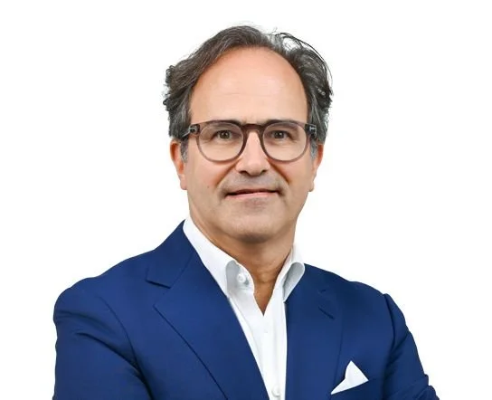 Ein Mann mit Brille, in einem blauen Anzug und weißem Hemd, steht vor weißem Hintergrund.