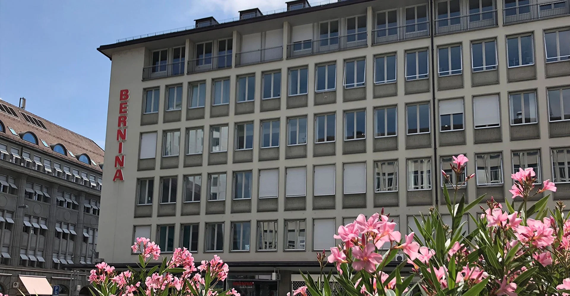 Mehrstöckiges Gebäude mit roten Buchstaben, die 'BEERNINA' lesen. Im Vordergrund pinkfarbene Blumen. Das Gebäude hat viele Fenster und Balkone, sonniger Tag, blauer Himmel.
