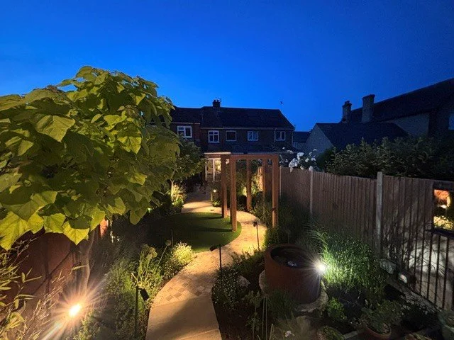 Nighttime view of a quaint backyard garden with a winding pathway, illuminated by outdoor lights, surrounded by lush plants and a wooden fence, with houses and a deep blue sky in the background.