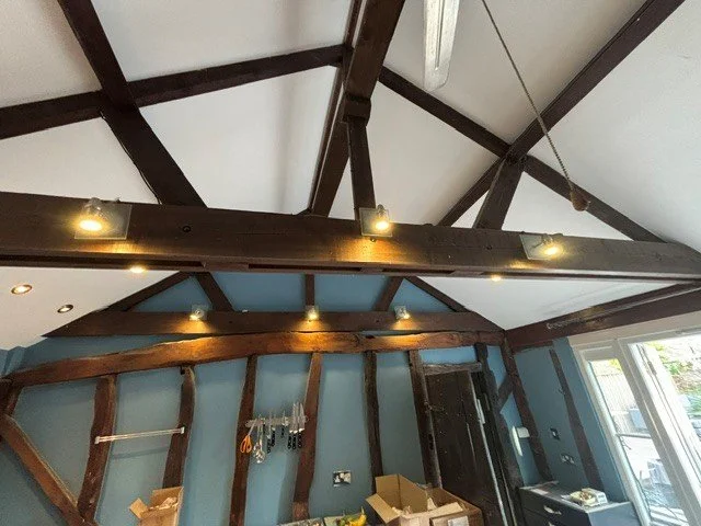 Interior of a room with exposed wooden beams on the ceiling and walls, ceiling lights, and a large window with a view outside.