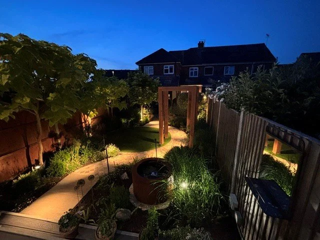 A backyard garden at night with a glowing pathway, plants, a wooden arch, and a fence, illuminated by small lights.
