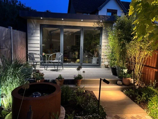 Nighttime backyard patio with outdoor seating and garden lighting, leading to a house with large sliding glass doors.