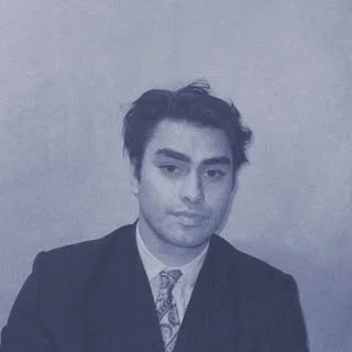 A young man with dark, styled hair wearing a suit and patterned tie, looking at the camera with a neutral expression.