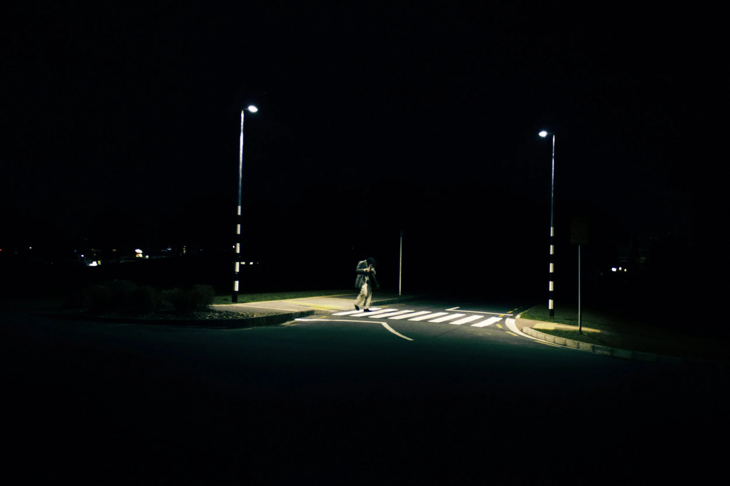 A person walking across a pedestrian crosswalk at night, illuminated by streetlights, in an empty parking lot or road area.