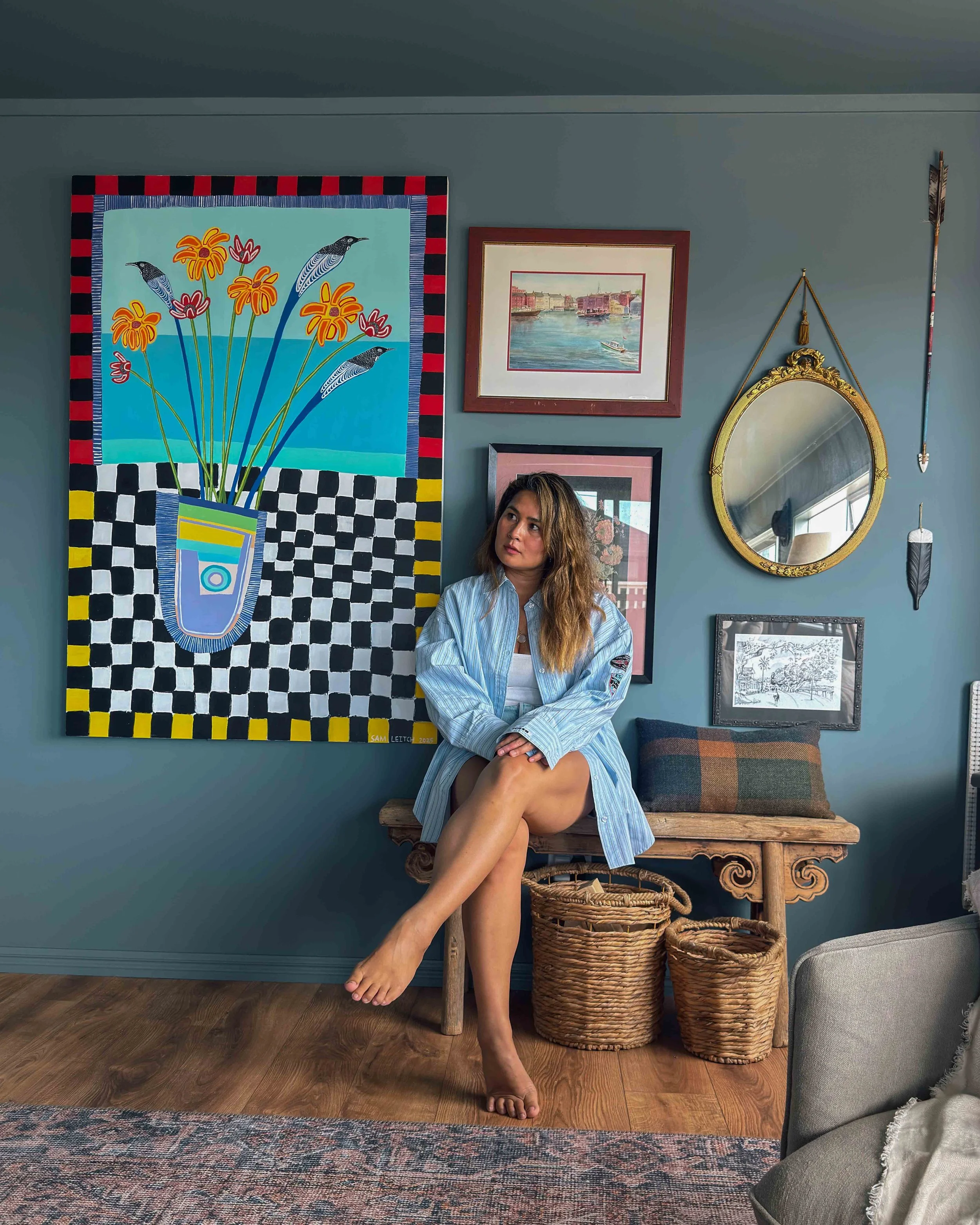Young woman with wavy hair sitting on a wooden bench with one leg crossed over the other, wearing a blue striped shirt and white top, in a room decorated with colorful artwork and vintage-style furnishings.