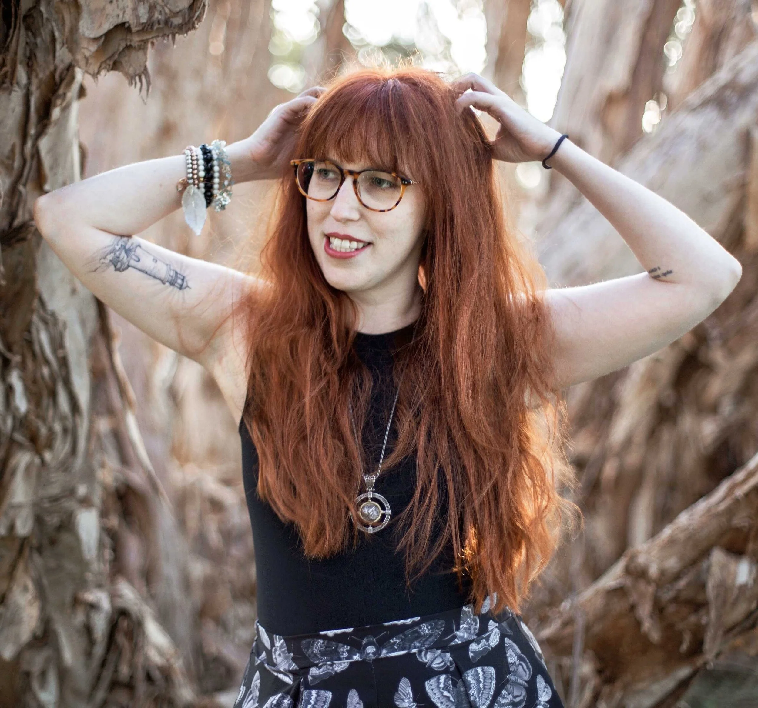 A white woman with long red hair and glasses stands somewhat dramatically in paperbark bushland.