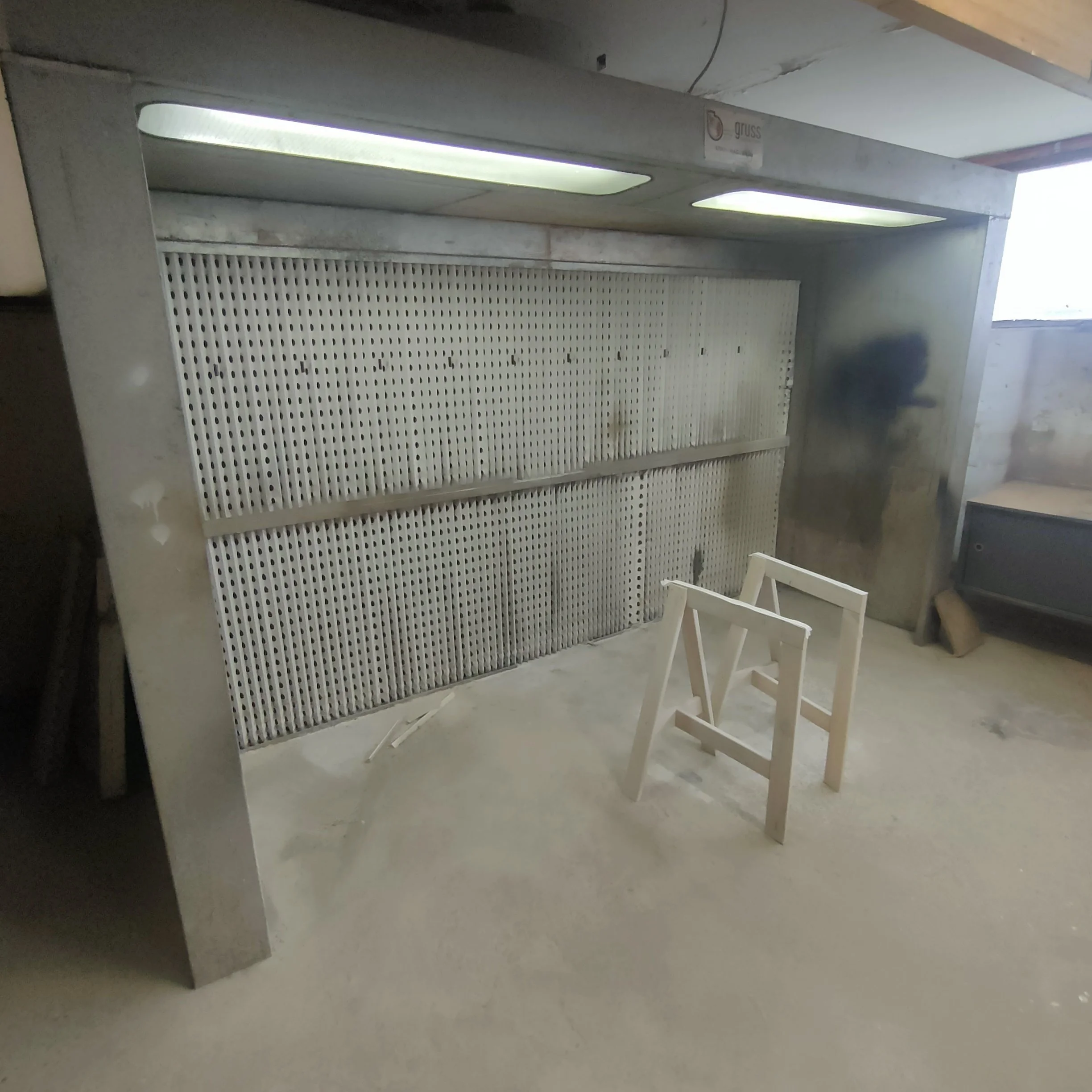 Un plan de travail ou comptoir en métal avec une grille perforée, une lumière fluorescente au plafond et deux chaises en bois blanc.