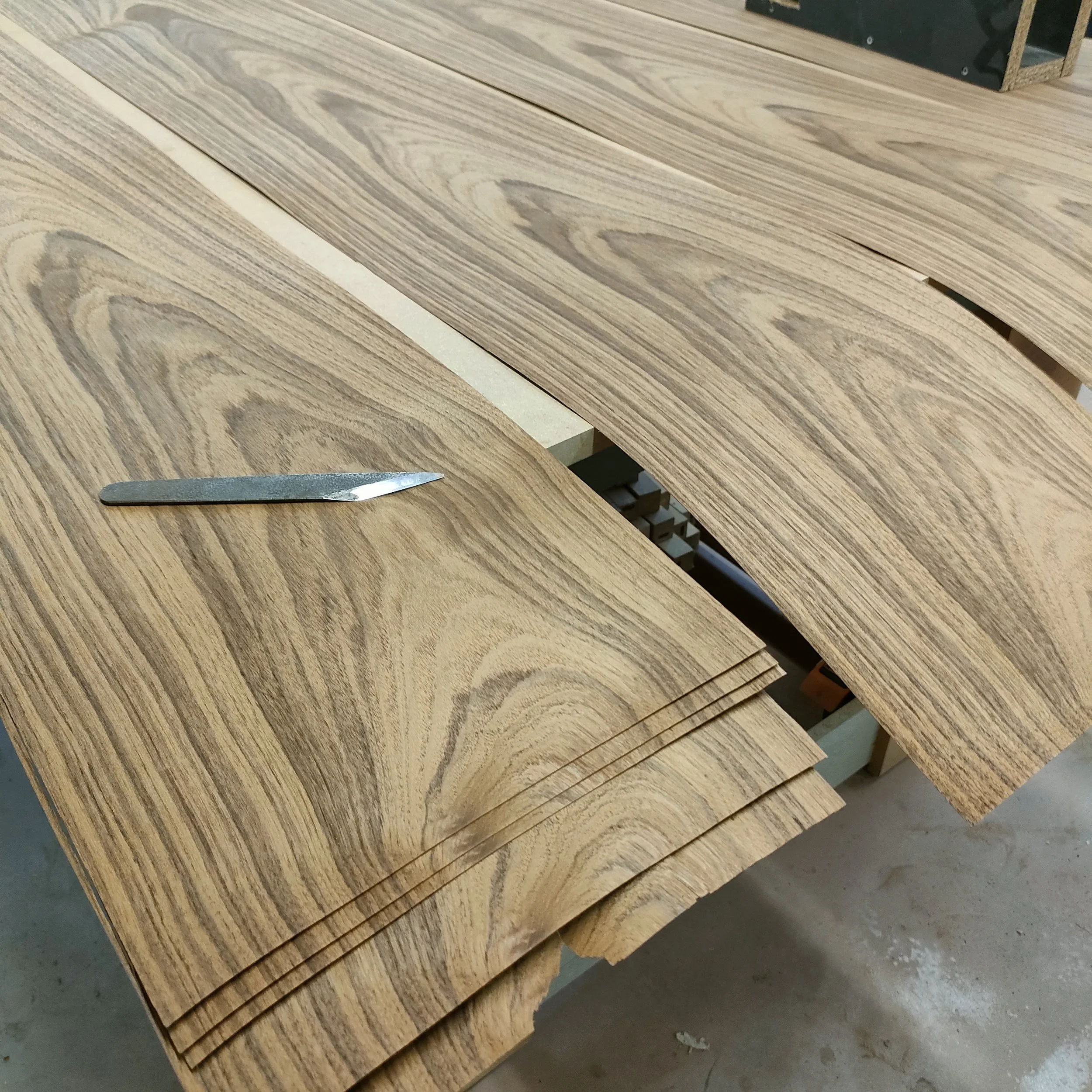 Planches de bois clair empilées sur un sol en béton, avec un couteau de menuisier posé au premier plan.
