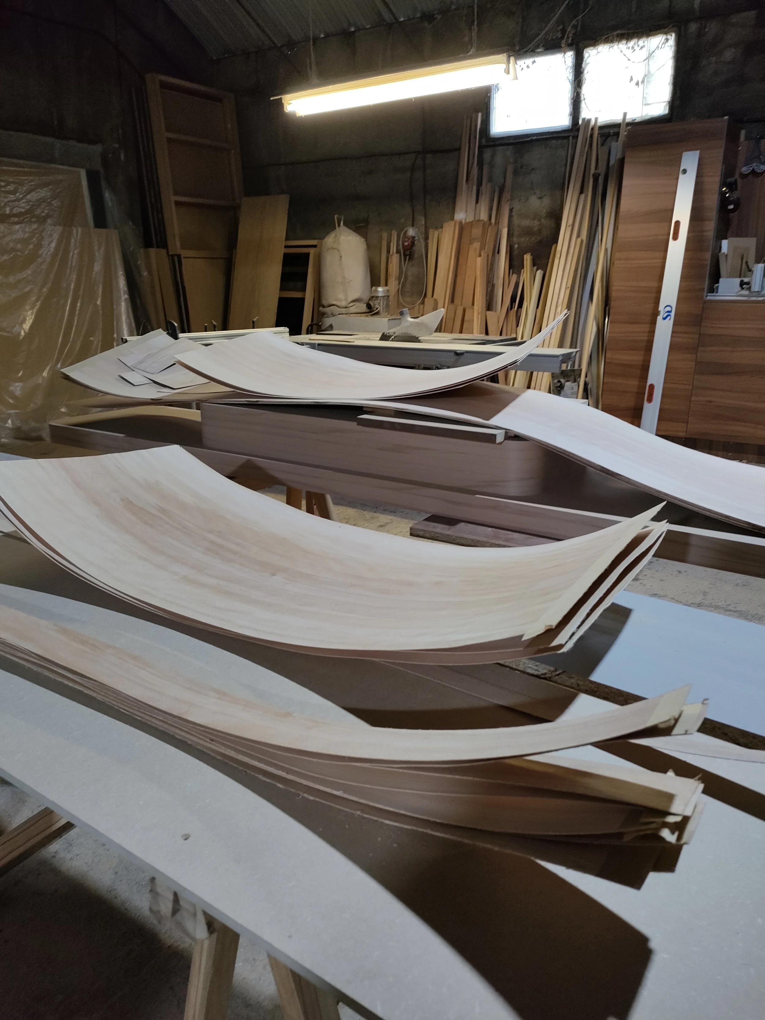 Un atelier de menuiserie avec des morceaux de bois courbés sur une table, des planches de bois empilées au mur et une règle de nivellement.