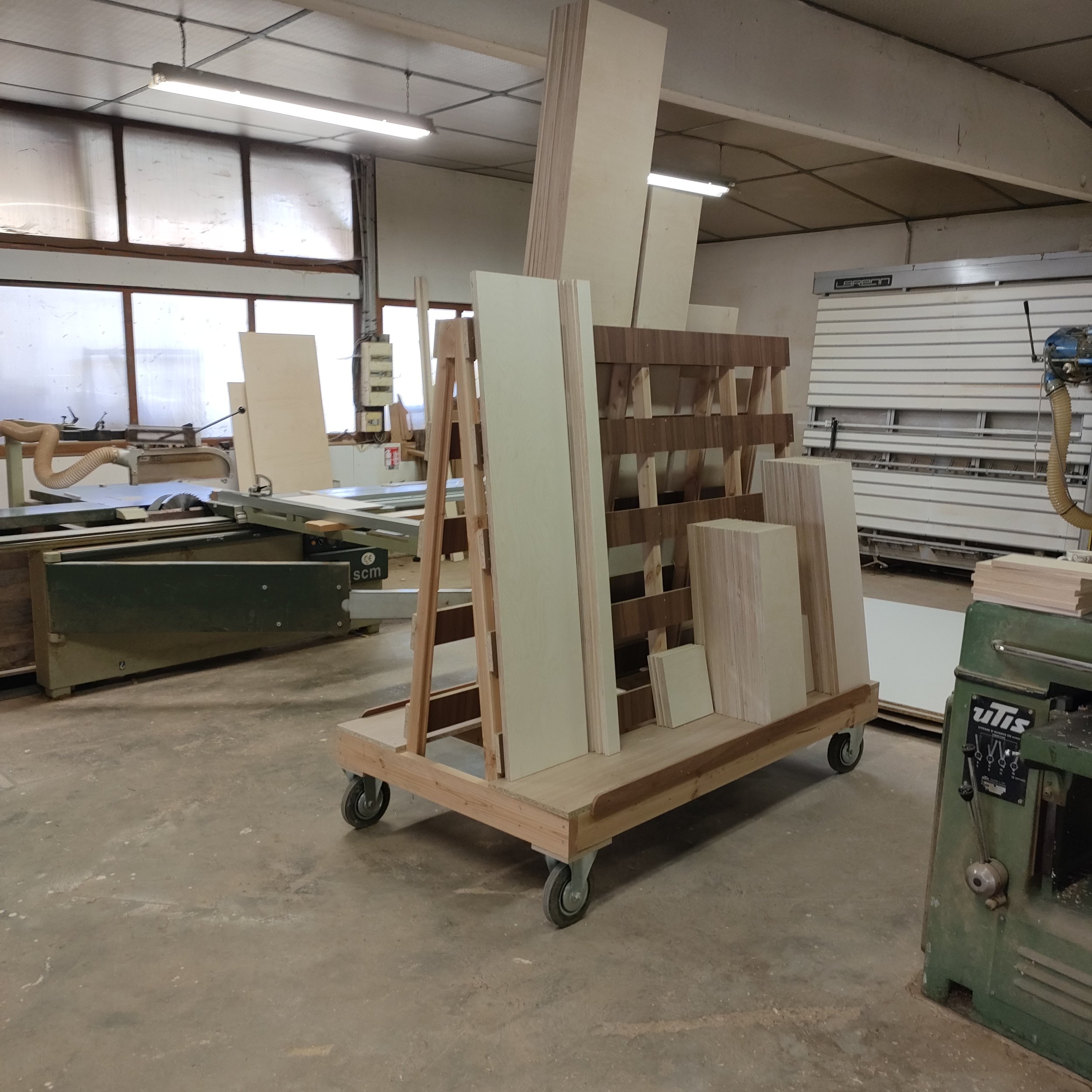 Un chariot en bois sur roues dans un atelier de menuiserie avec des étagères contenant des planches en bois et des outils, éclairé par des luminaires suspendus.