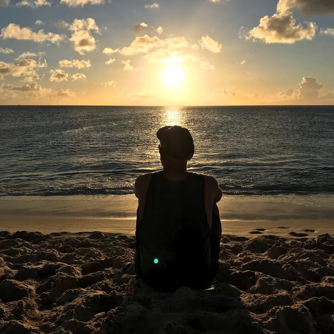 Une personne assise sur le sable regarde le coucher de soleil au-dessus de l'océan, avec quelques nuages dans le ciel.