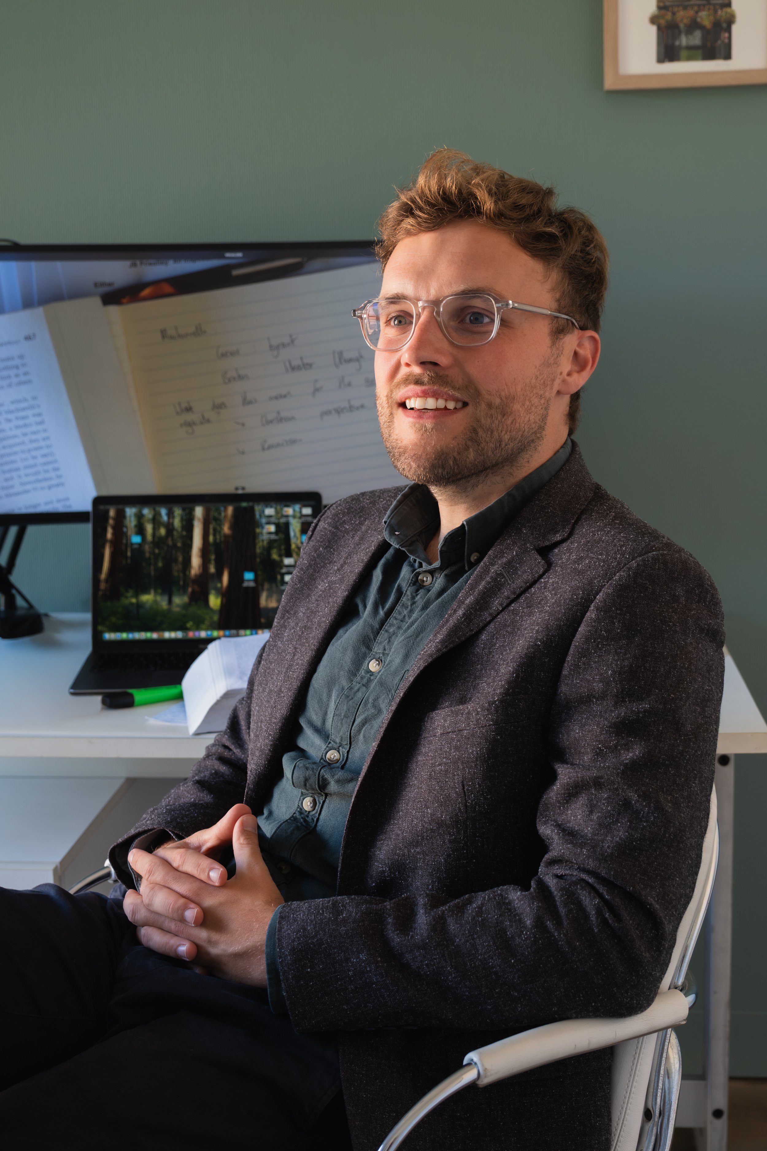 A man with glasses and a beard, wearing a gray blazer and black shirt, sitting in an office. Behind him is a computer monitor, a notepad with handwritten notes, a green marker, and a laptop displaying a forest scene on the screen.