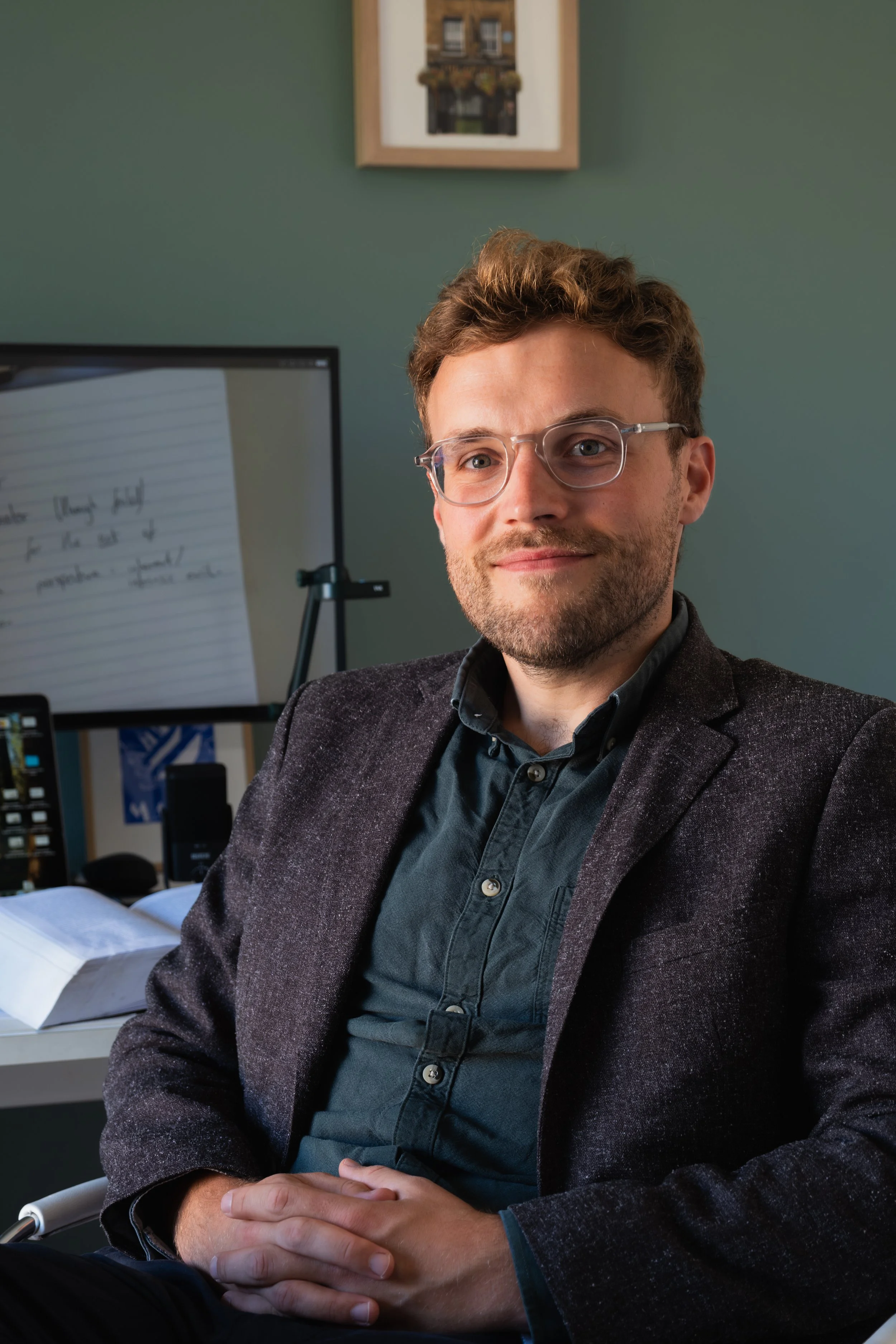 A man with glasses and a beard sitting at a desk with open notebooks, a computer monitor, and a phone in the background, in a room with a green wall and a framed picture.