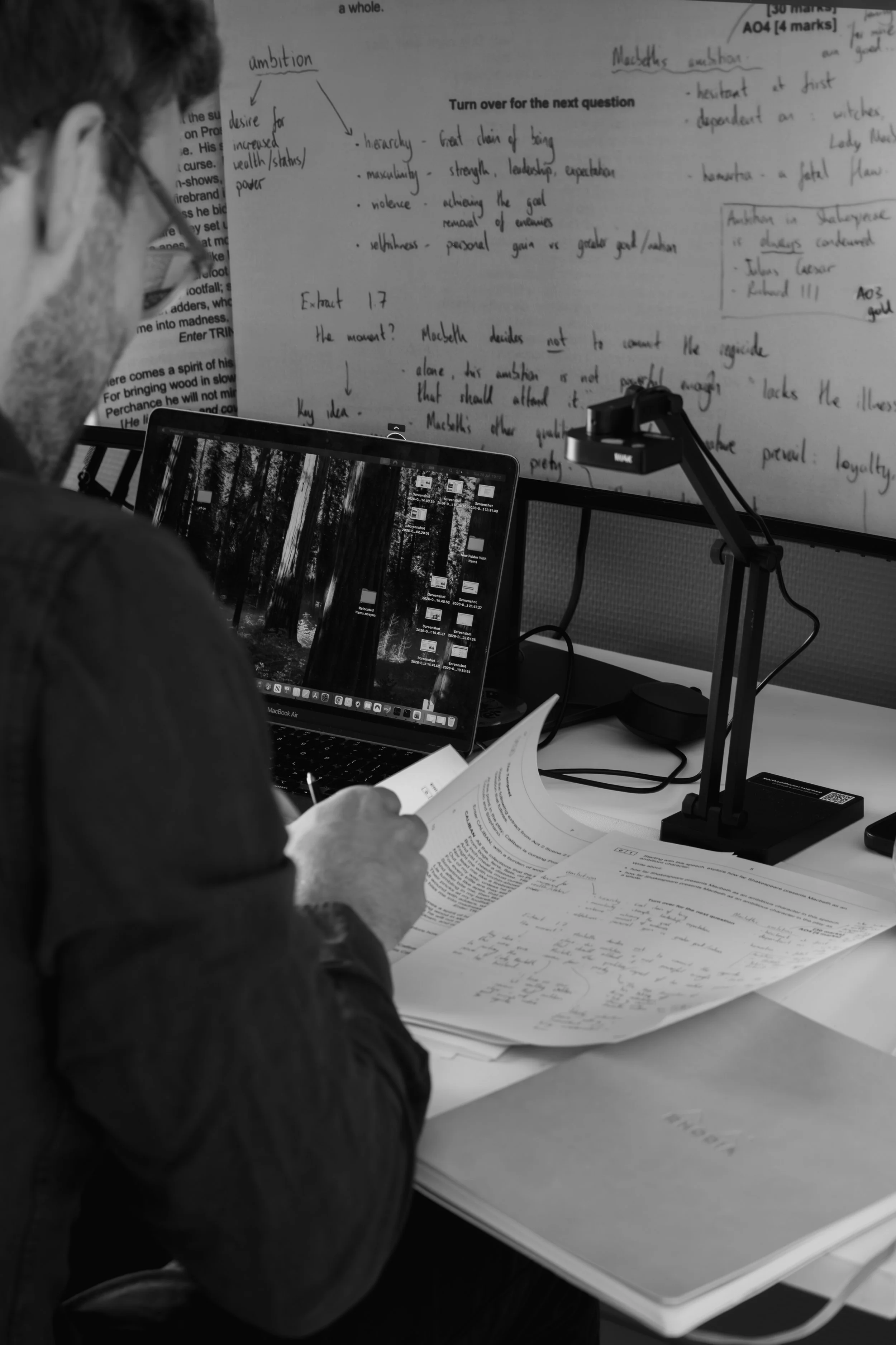 A person with glasses working at a desk with papers, reading a document, with a laptop open displaying a forest scene, and notes pinned on a board behind them.