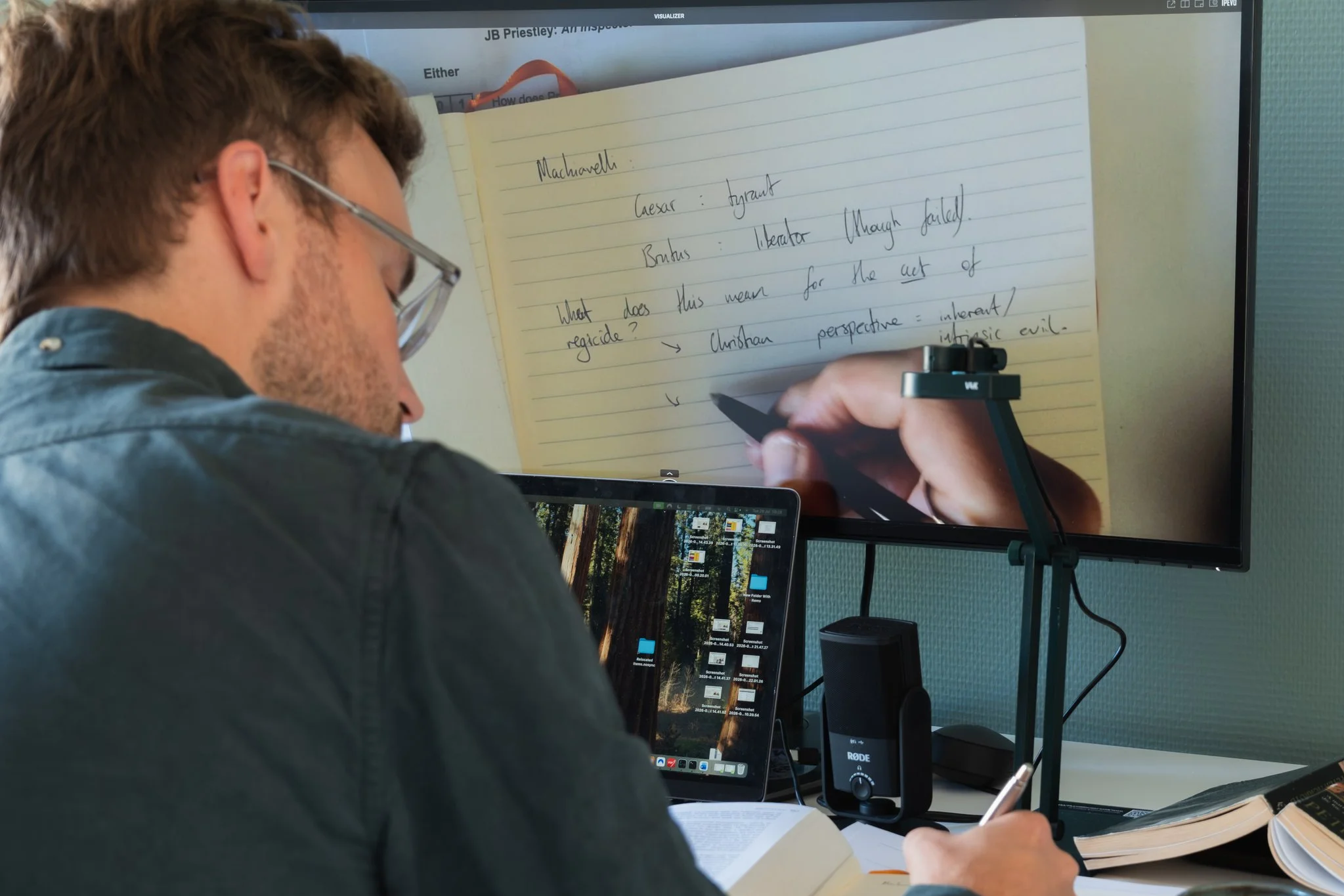 Alex is working at a desk with a computer monitor and a laptop, writing notes. The computer monitor displays a handwritten note on a yellow notepad. The desk has books, papers, and a microphone.