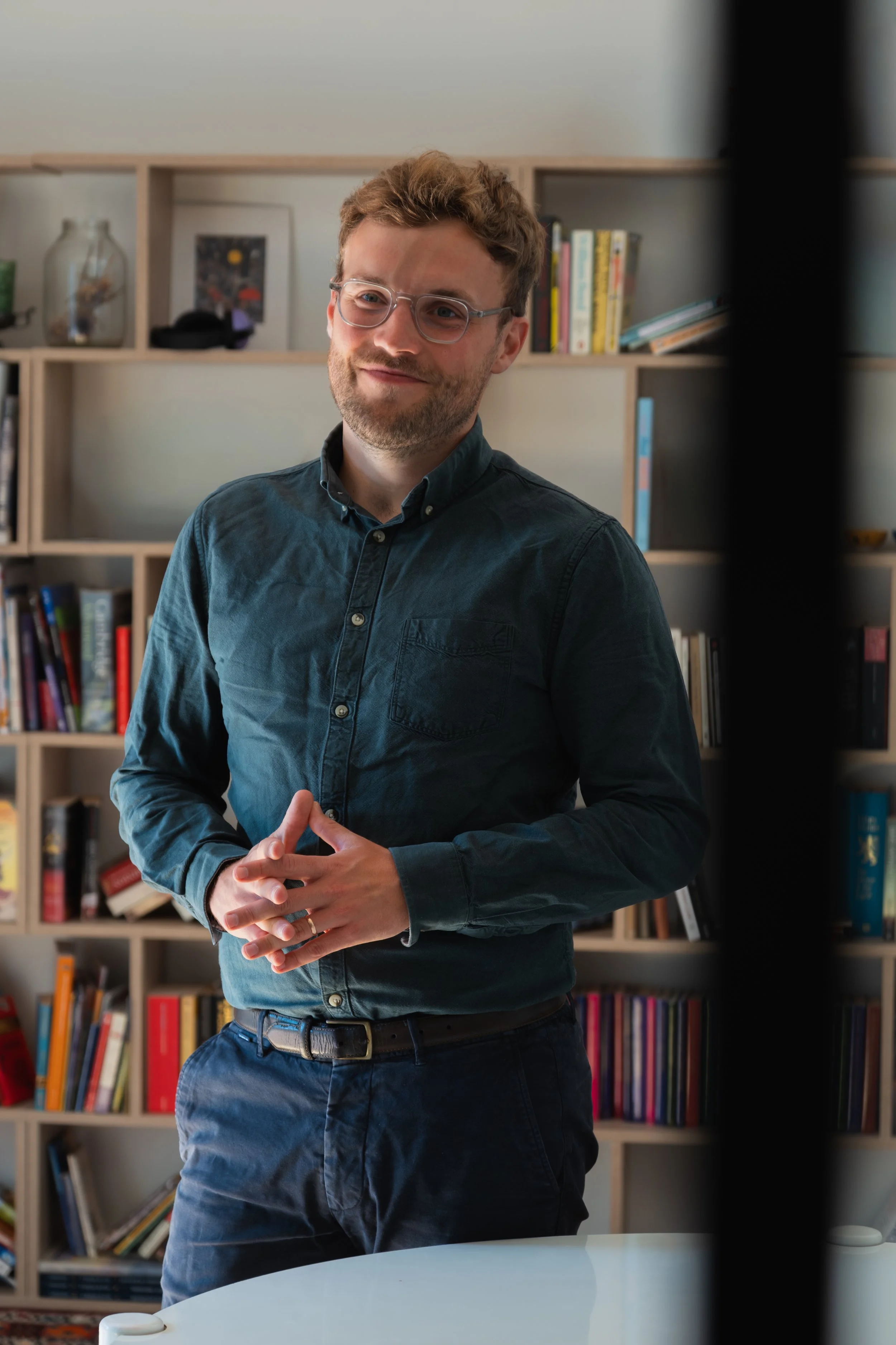 A man with glasses wearing a dark button-up shirt standing in front of a bookshelf filled with books and a framed picture, with his hands clasped in front of him.