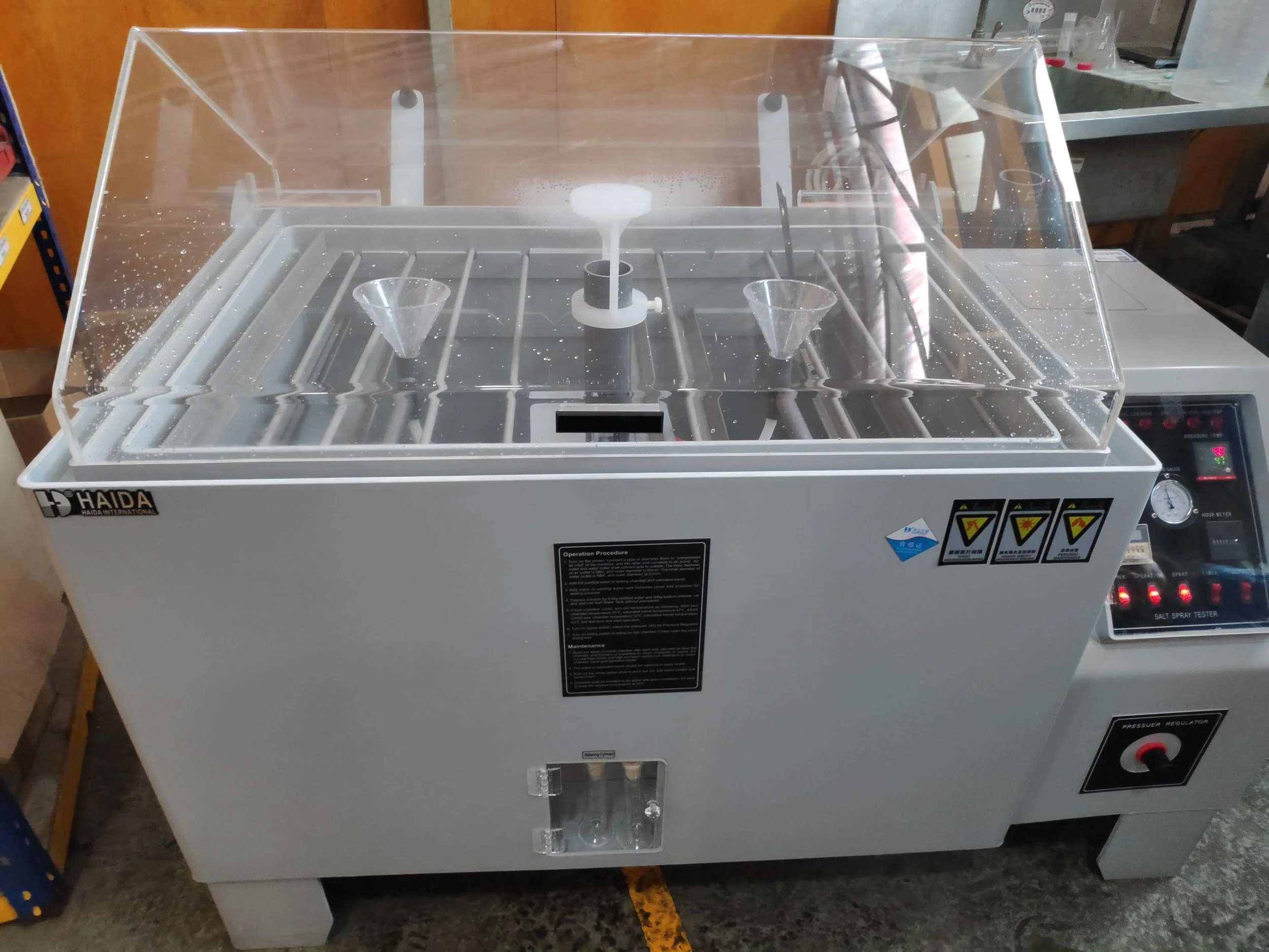 A laboratory salt spray testing chamber with a transparent plastic cover, four funnel-shaped containers, and control panels on the front