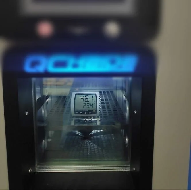 A climate chamber has a glass door showing its interior, where a fan and internal components are visible.