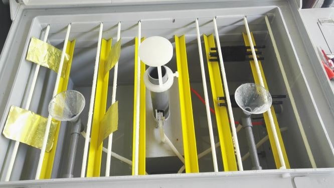 Salt Spray Chamber setup with yellow and white barriers, plastic containers, and metal rods inside a glass case.