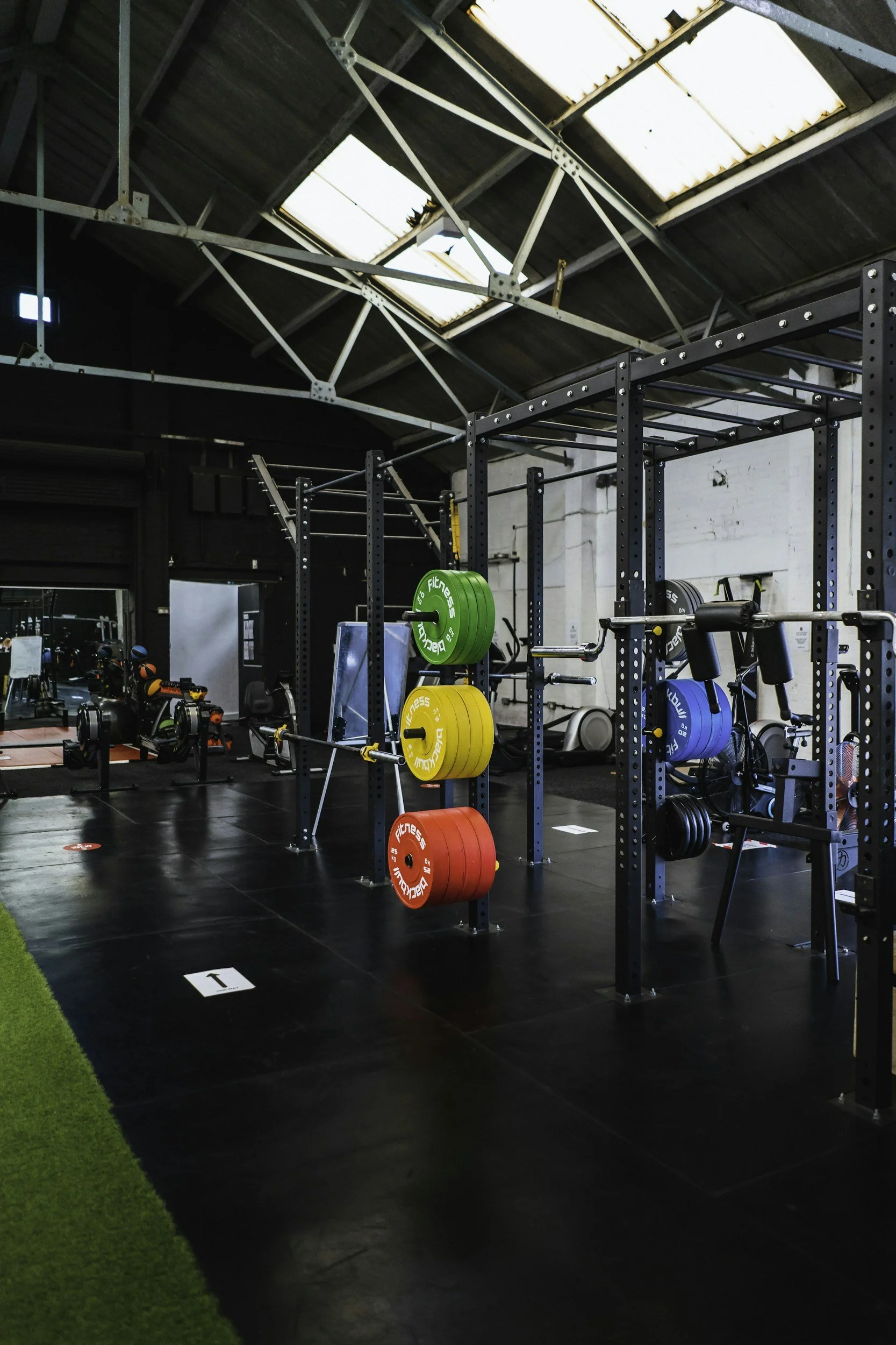 Interior view of a gym with black flooring, weightlifting equipment, colorful weight plates, rowing machines, and cutting-edge fitness machines. The gym has an industrial ceiling with skylights, a mix of black and white walls, and a section of artificial green turf.