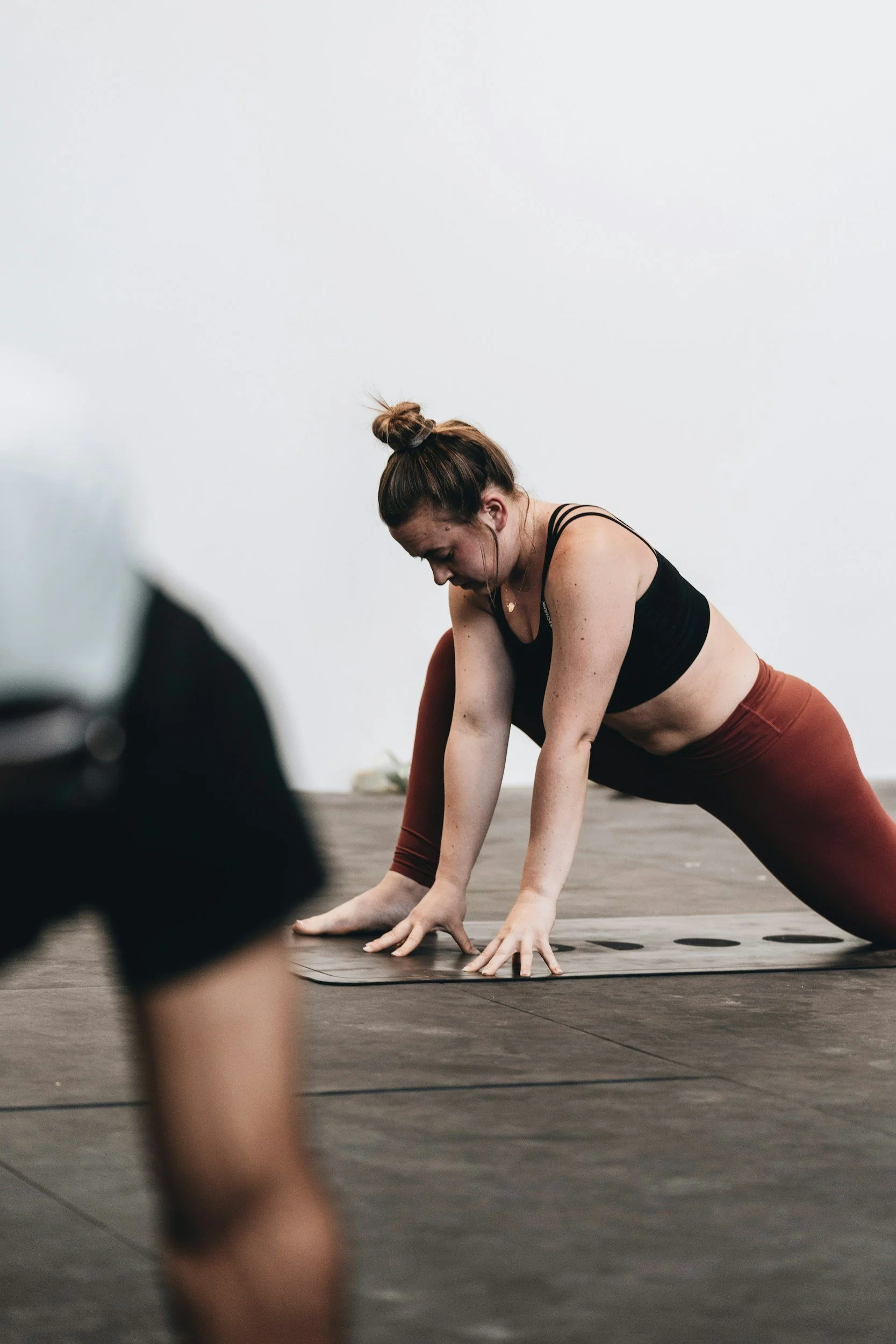 A woman with red hair in a bun is practicing yoga in a studio, wearing a black sports bra and red leggings, as she stretches on a yoga mat.