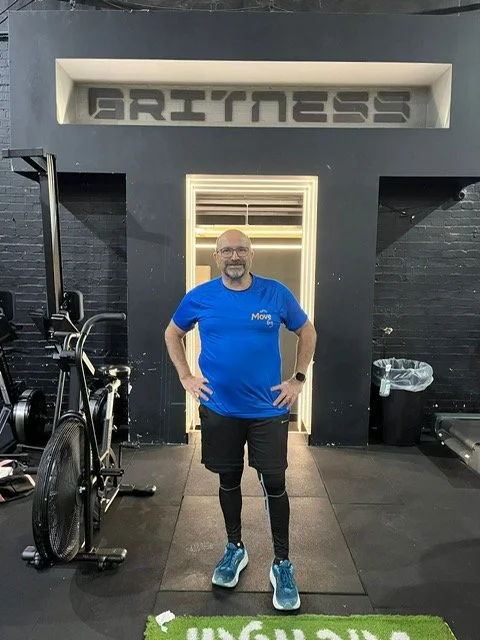 Man standing in a gym, posing for a photo, with the word 'BRYMESS' on the wall above him, wearing a blue t-shirt, black shorts, and athletic shoes.