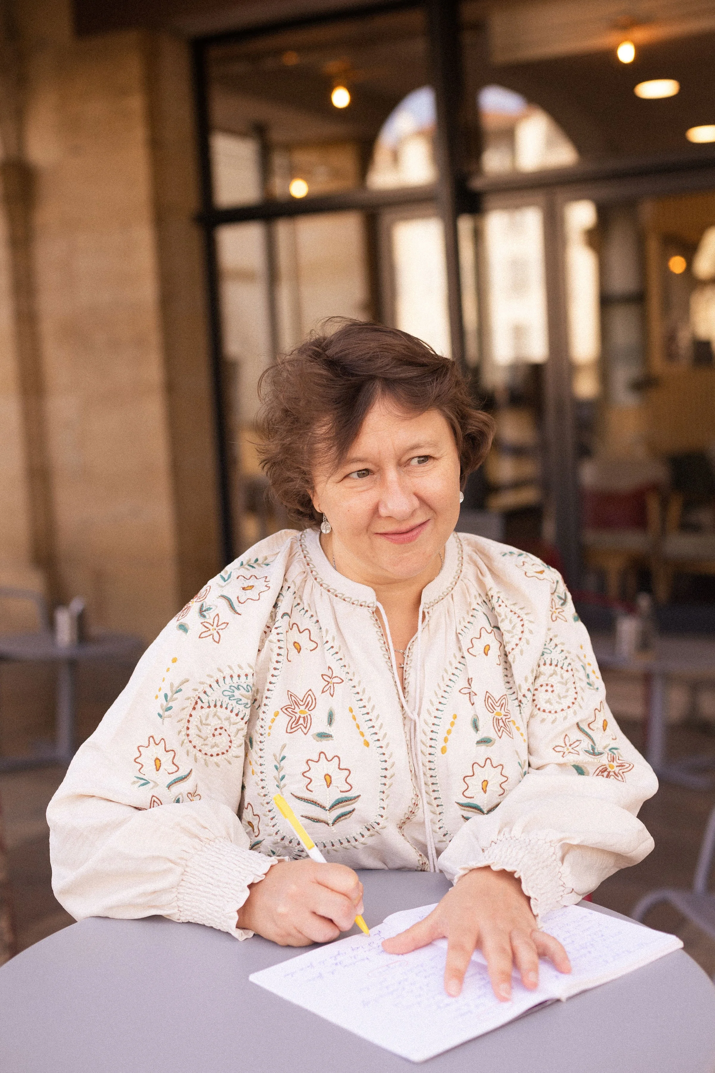 Une femme à cheveux courts, portant un vêtement blanc avec des motifs colorés, est assise à une table, écrivant dans un carnet avec un stylo jaune, dans un lieu lumineux avec de grandes fenêtres en arrière-plan.