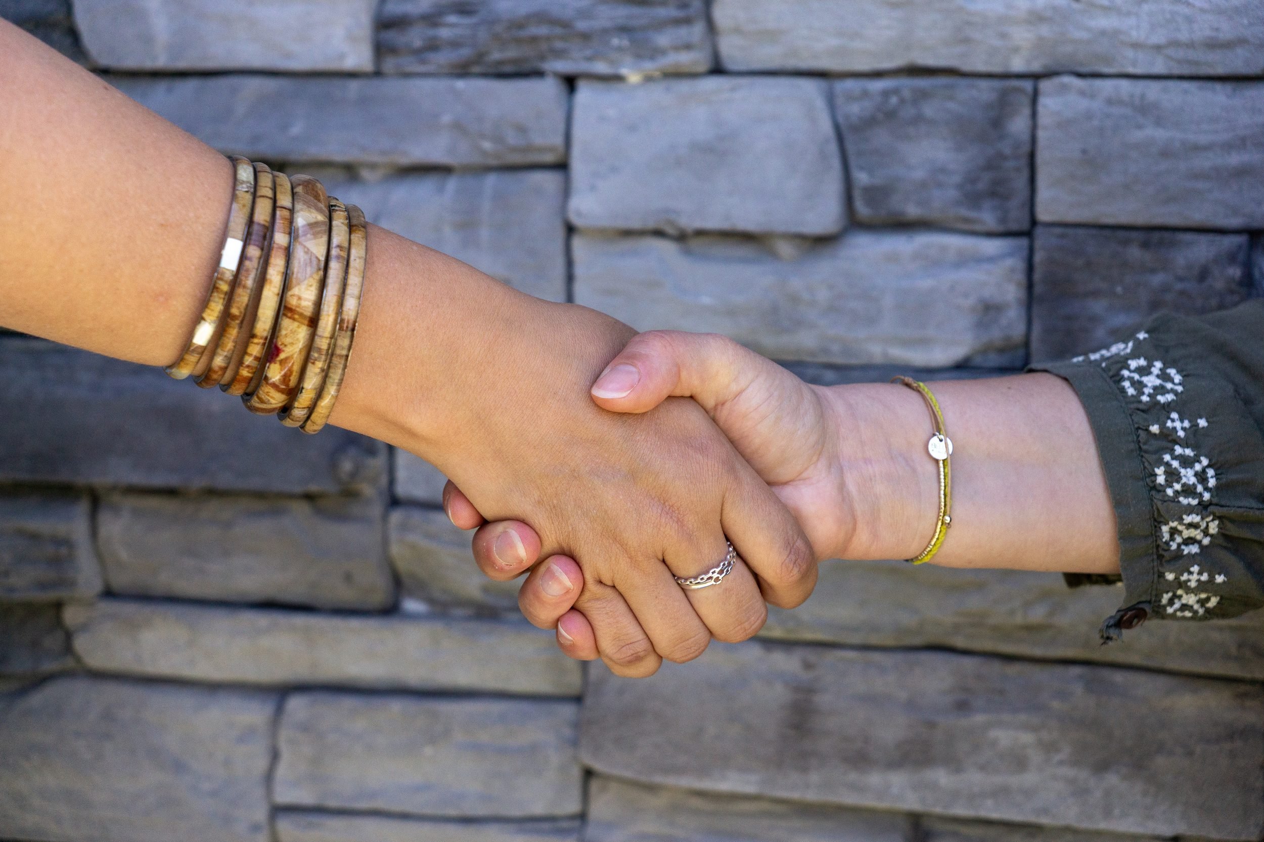 Deux personnes se tiennent par la main, portant chacune plusieurs bracelets et bagues. L'une porte une manche brodée et l'autre une bandoulière. En arrière-plan, un mur en pierres grises.