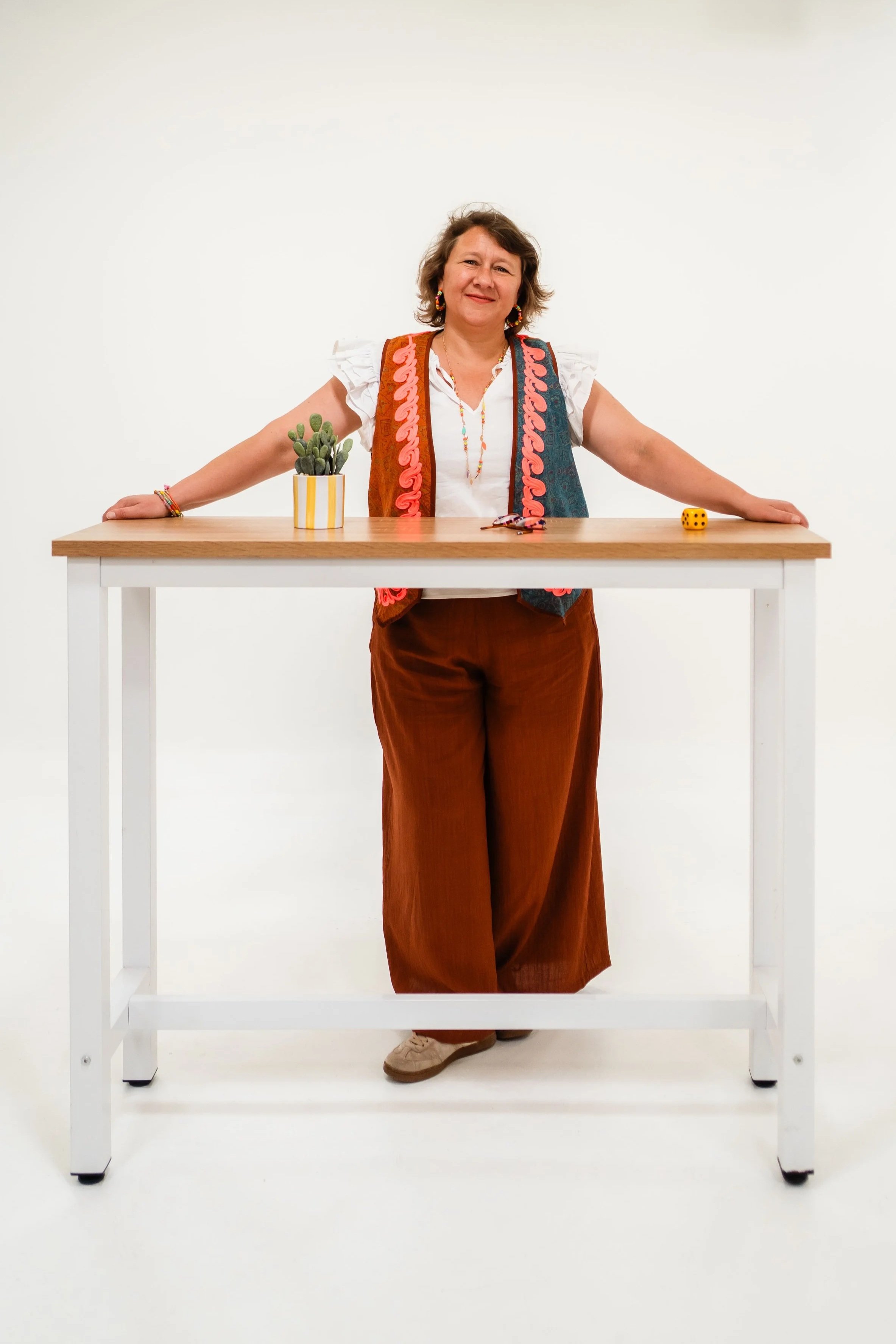 Femme debout derrière une table en bois, décorée d'une plante en pot, d'une paire de lunettes et d'un dé. Elle porte une veste colorée, une chemise blanche, un pantalon marron et des chaussures beiges, avec des boucles d'oreilles et un collier coloré.