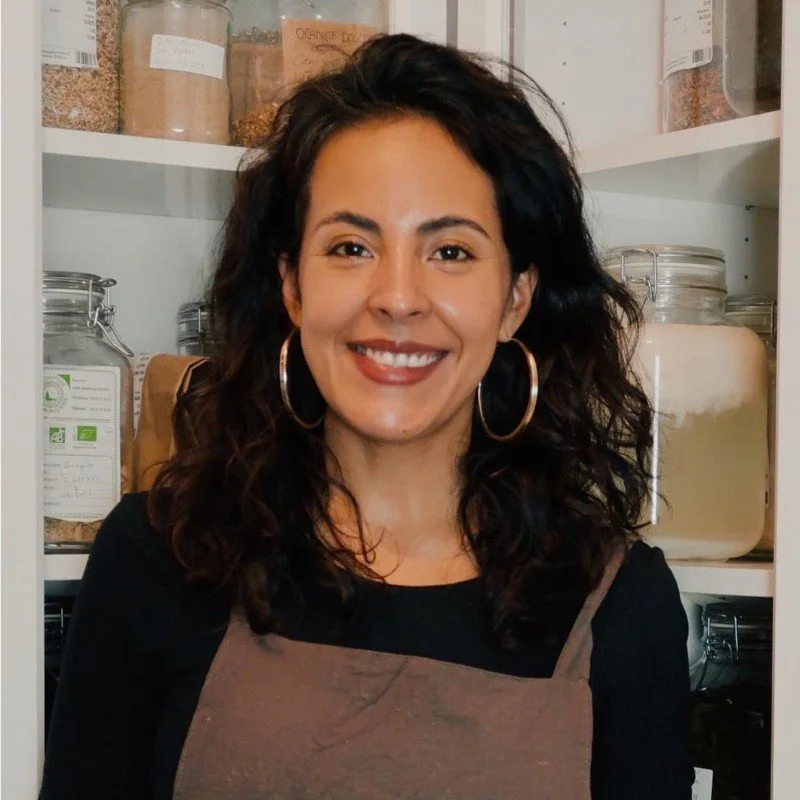 Une femme souriante avec de longs cheveux bouclés, portant de grosses boucles d'oreilles, dans une cuisine ou un espace de stockage avec des bocaux en verre contenant des épices ou des ingrédients alimentaires.