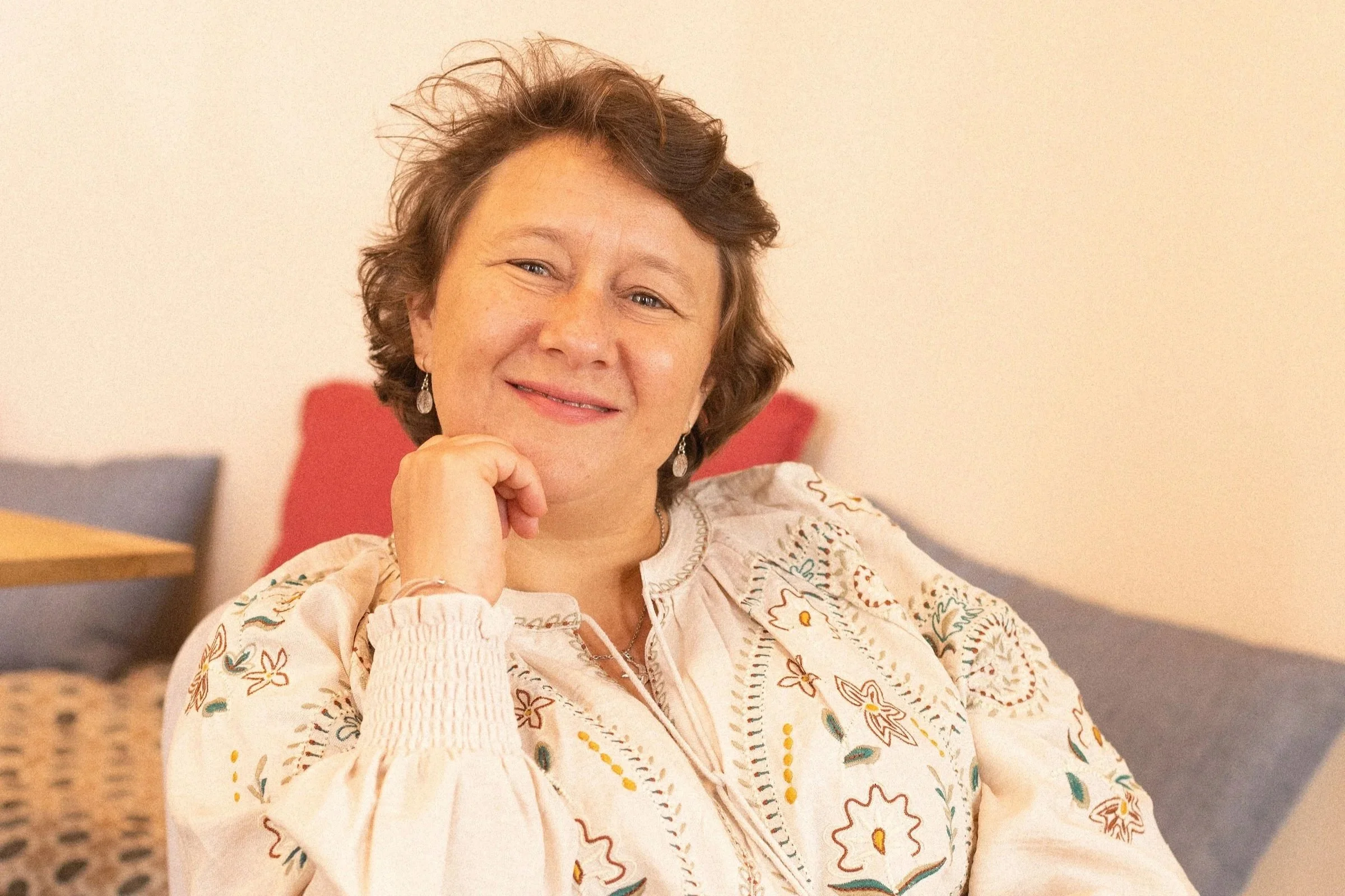 Une femme souriante avec des boucles brunes, portant des boucles d'oreilles en argent, est assise dans une pièce avec un fauteuil rouge en arrière-plan. Elle porte un haut à manches longues avec un motif floral.