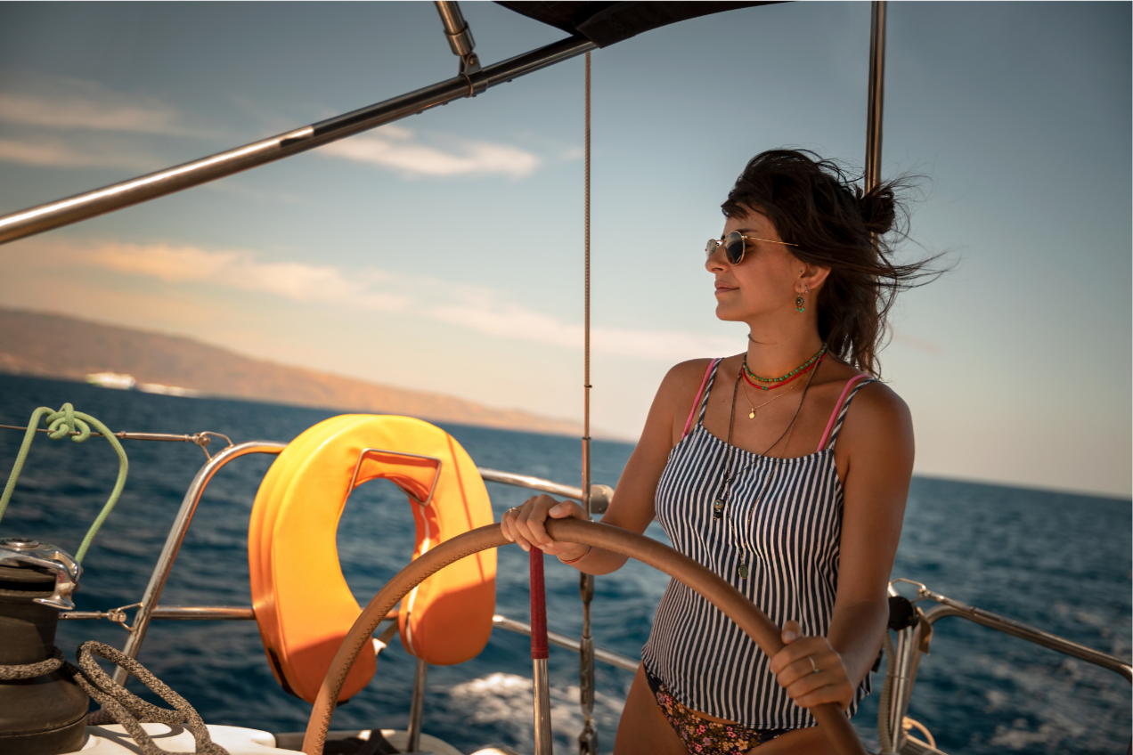 Une femme portant des lunettes de soleil, un maillot rayé et des bijoux, tient le gouvernail d'un bateau en mer lors d'une journée ensoleillée.
