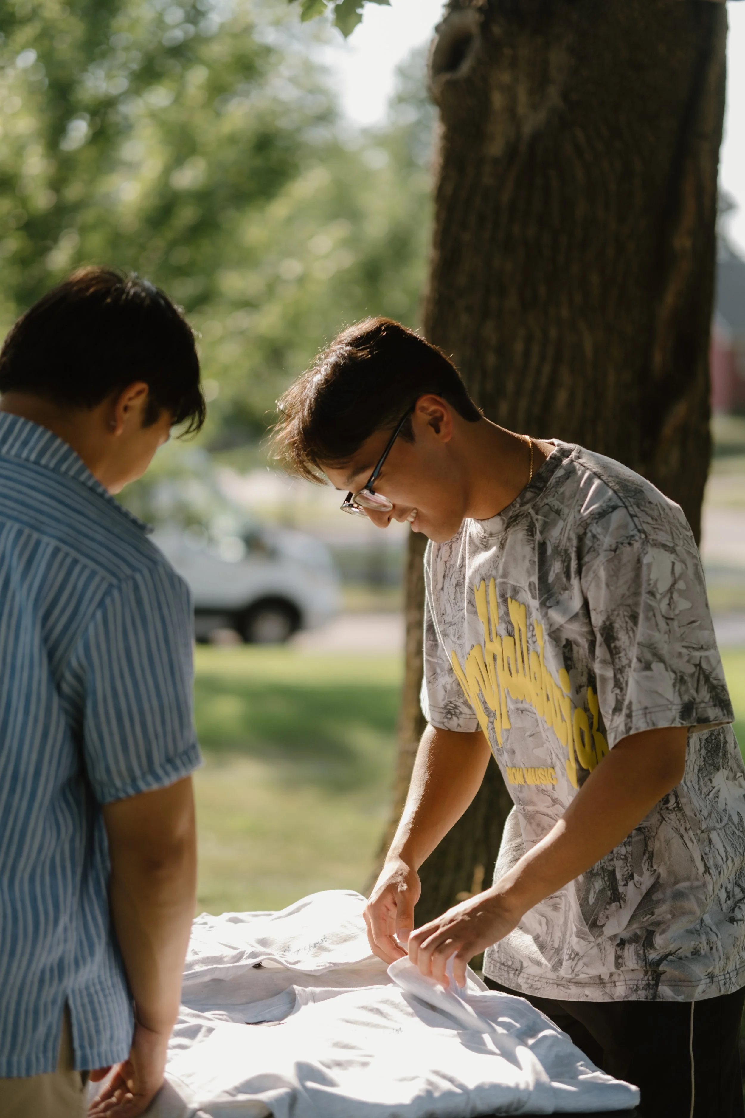 Two young men are standing outdoors near a tree, carefully handling a white cloth or sheet on a table. Merch. Jesus clothes. Faith apparel.