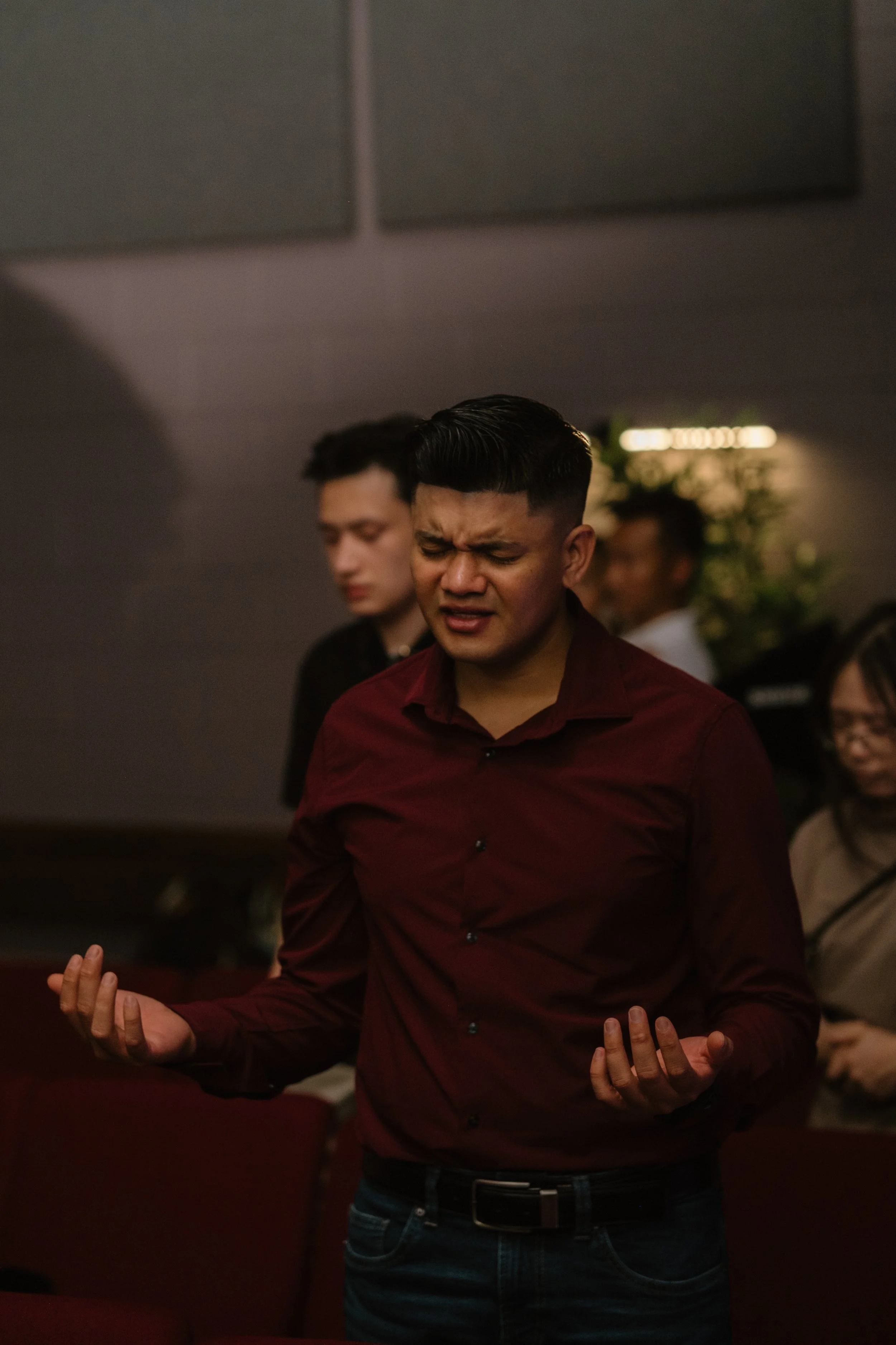 A young man dressed in a maroon shirt and jeans is standing with eyes closed, hands raised, and a facial expression of concentration or prayer, in a dimly lit room with other people standing behind him.