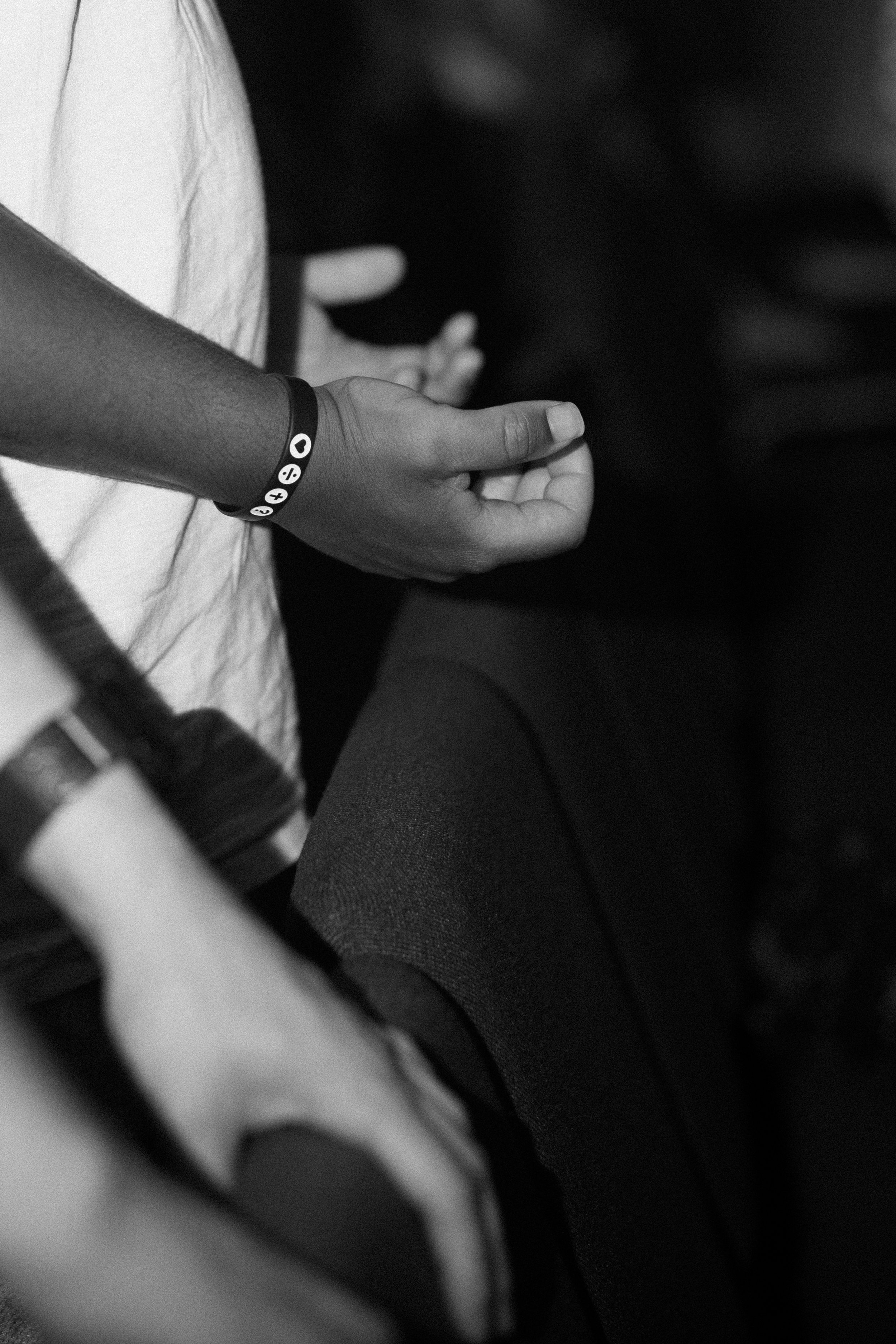 Close-up of a person's hand and wrist, wearing a bracelet with various symbols, resting on their leg, with blurred background elements. Worship hands. Deep worship. Moving worship. Good worship. Modern worship.