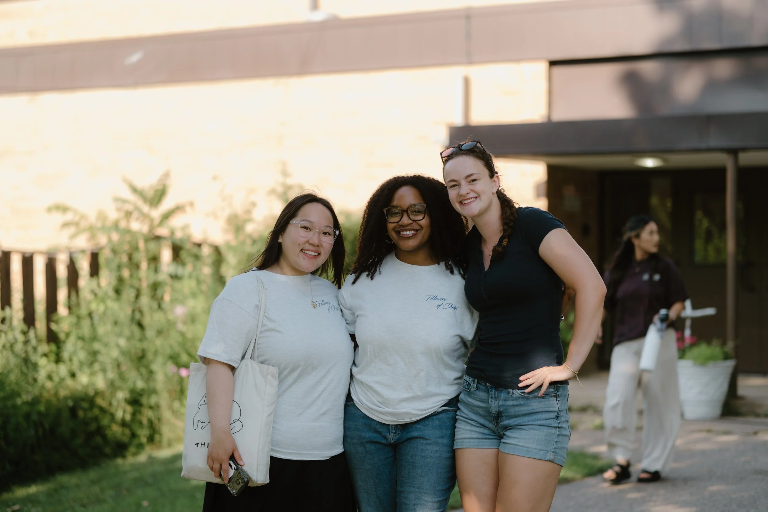 Three young women smiling and standing close together outdoors on a sunny day.