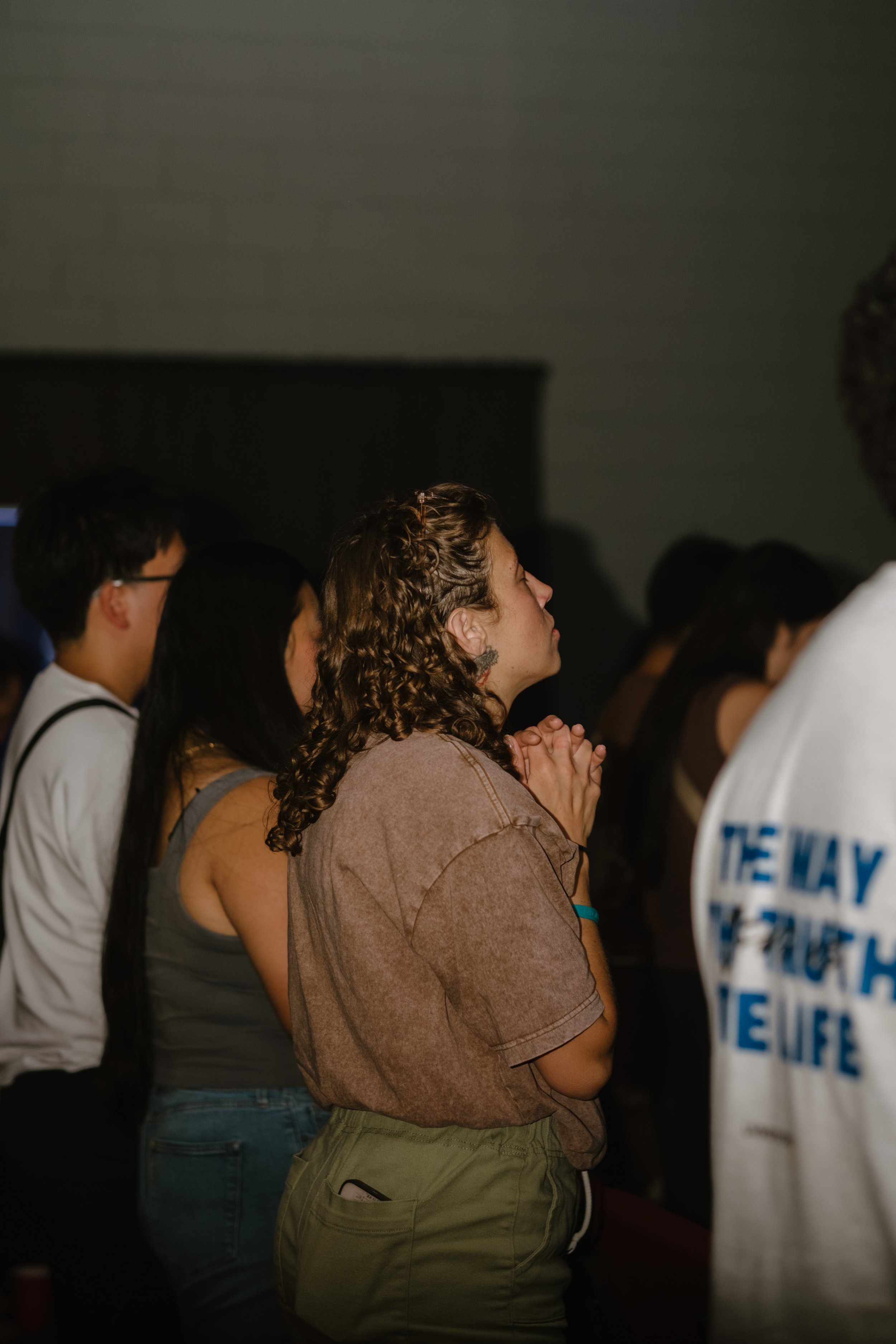 People standing with hands clasped, eyes closed, appearing to be praying or meditating in a dark indoor setting.