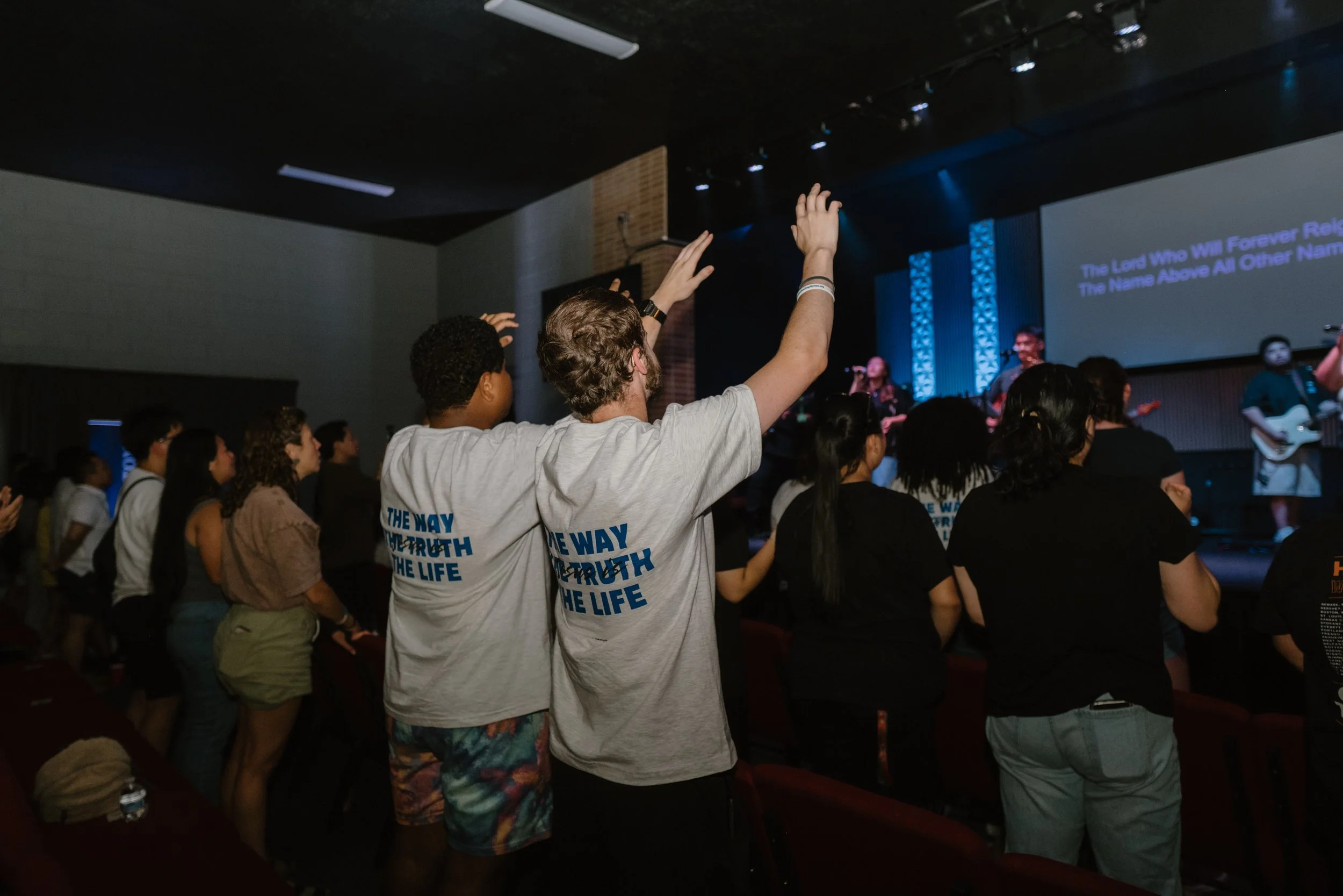 People worshipping and raising hands during a church service with a live band performing on stage, and lyrics projected on a screen behind them.