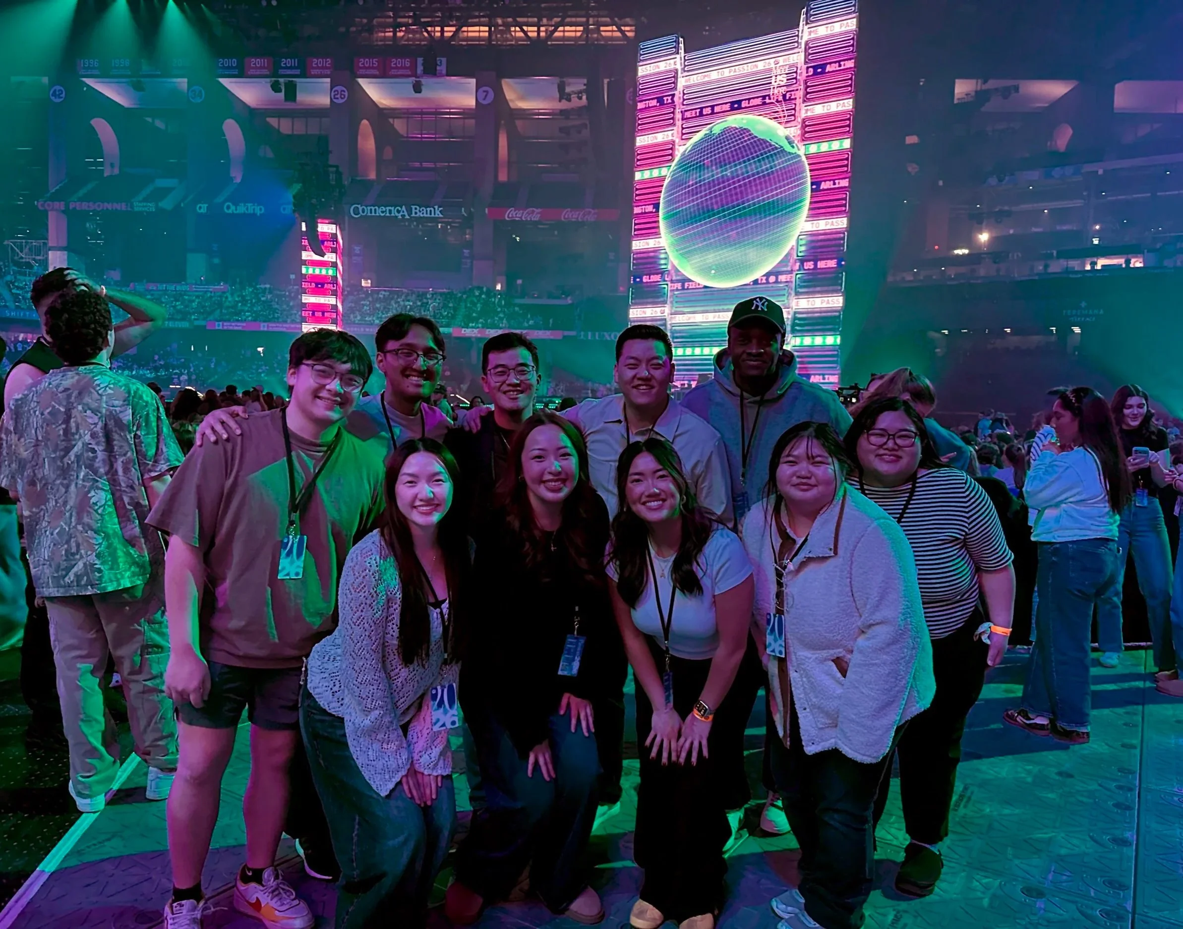 A group of young people smiling and posing for a photo at an indoor event or concert. The background features large LED screens with vibrant graphics and text. Church conference. Church gathering. Young adult gathering. YA night