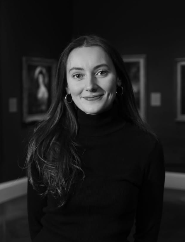 A young woman with long hair wearing a black turtleneck and hoop earrings, smiling in an art gallery.