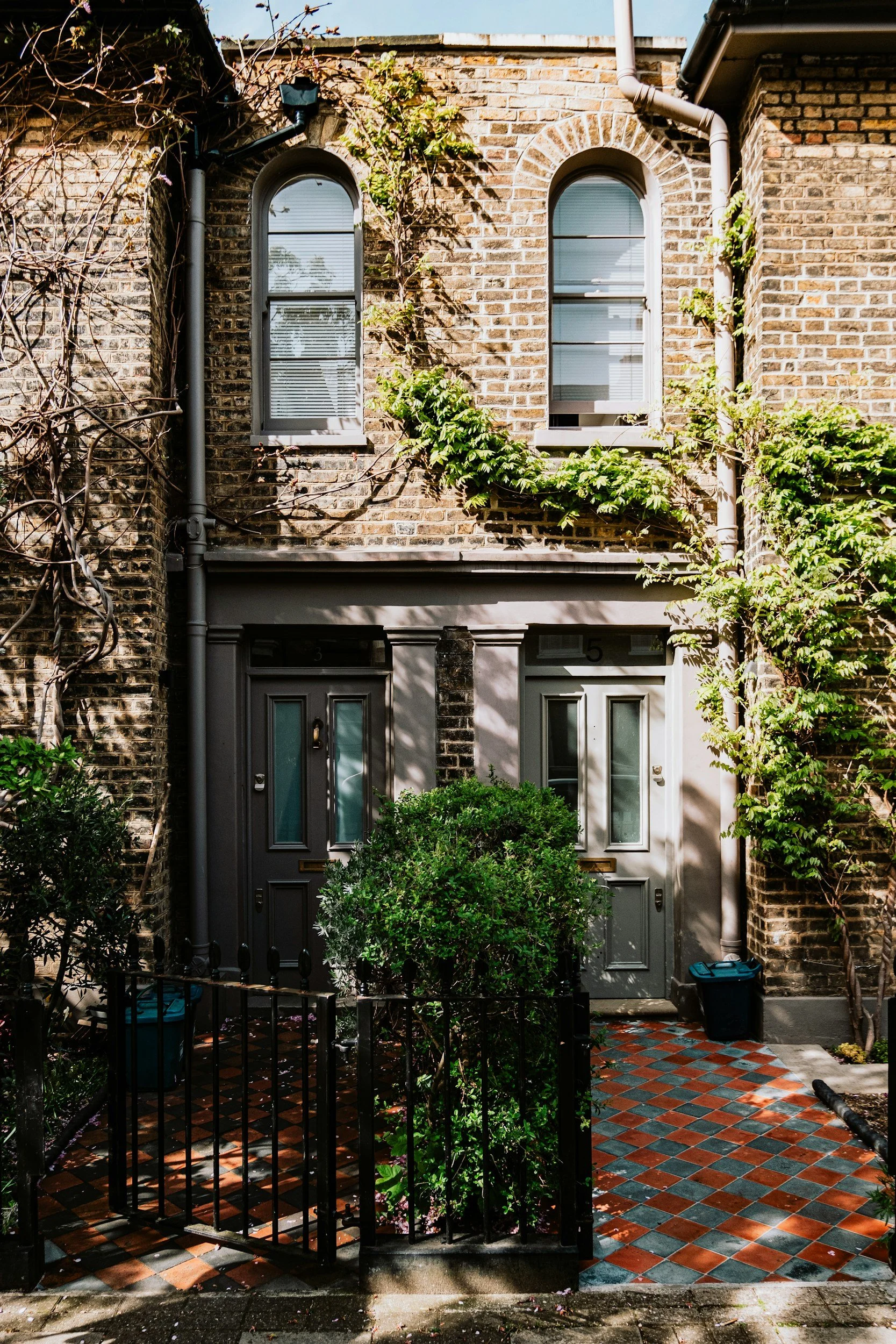 Front view of a brick townhouse with two arched windows on the second floor. The entrance has double gray doors with glass panels, a small front garden enclosed by a black metal fence, and a red and black checkered tile pathway. There are leafy plants and vines growing on the facade, and a gutter pipe on the right side.
