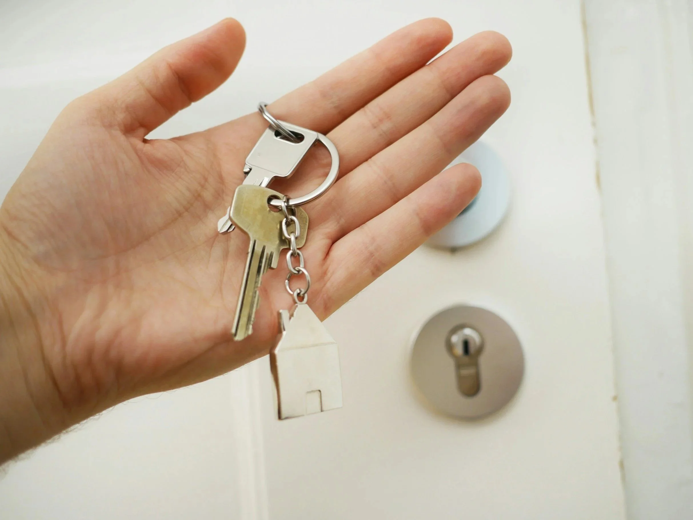 Close-up of a hand holding a set of keys with a keychain, near a light switch with an outlet on a light-colored wall.