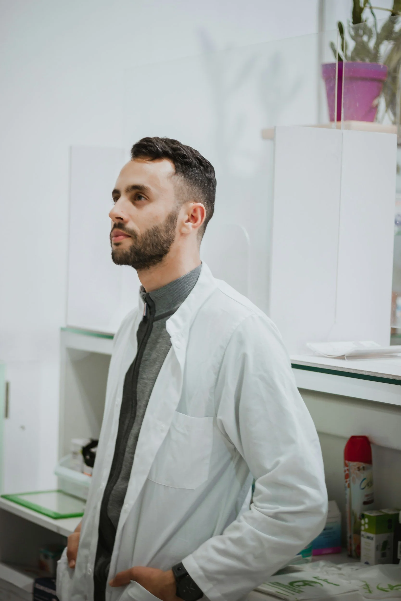 A man with short dark hair, a beard, and light skin wearing a gray shirt and white lab coat stands in a laboratory. Behind him are shelves with various supplies and a purple potted plant on top.