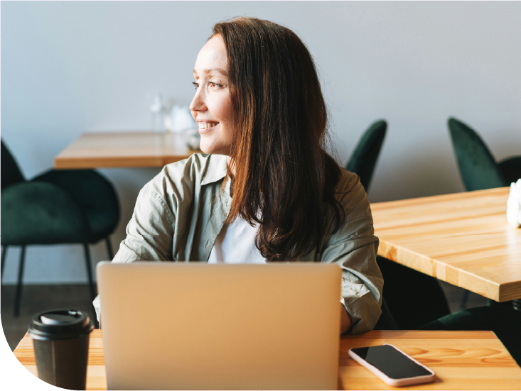 A woman with long brown hair sitting at a wooden table with a laptop, a coffee cup, and a smartphone, smiling while looking to her left in a modern, lightly decorated cafe or co-working space.