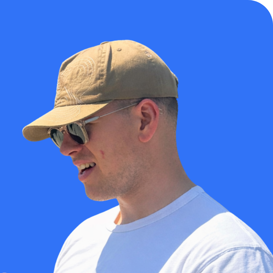 Profile of a smiling young man wearing sunglasses, a beige cap, and a white t-shirt against a bright blue background.