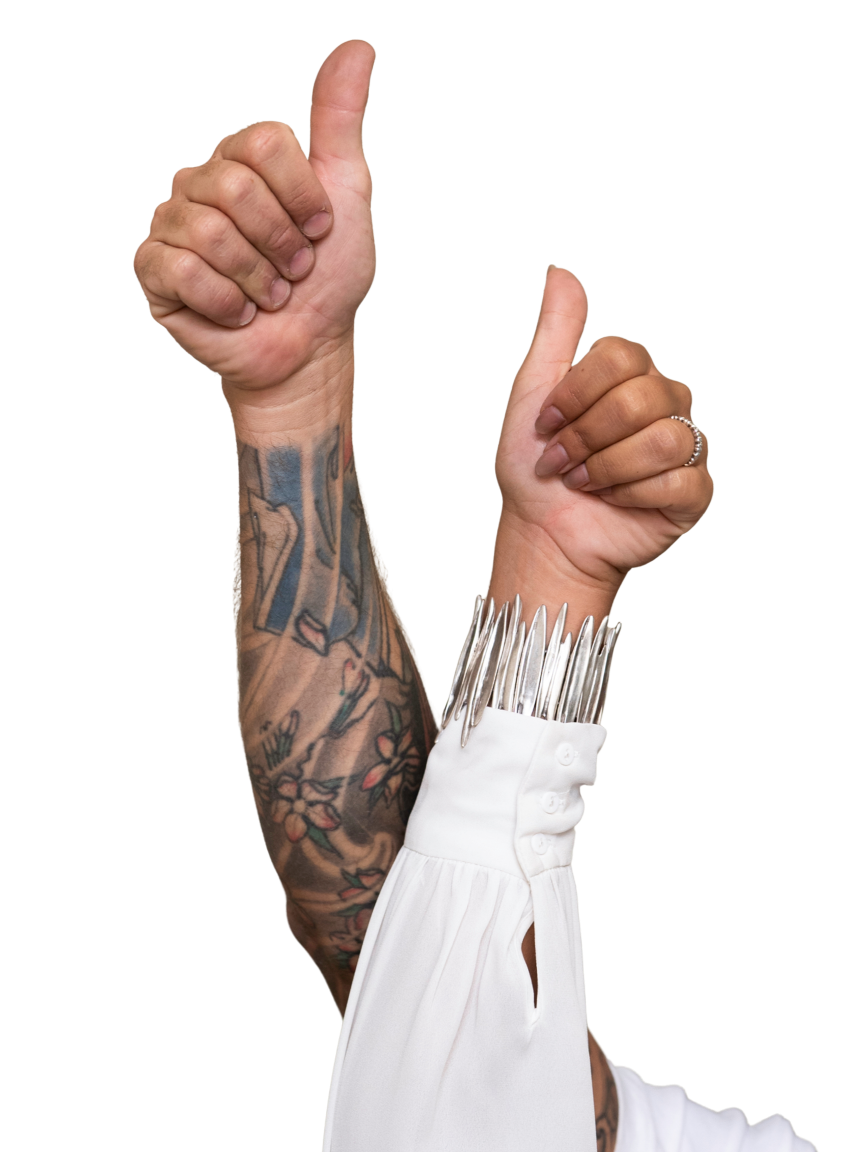 A person with tattooed arm and a ring on finger is wearing a white long sleeve shirt with metallic spiked cuffs, holding up thumbs in a gesture of approval against a black background.