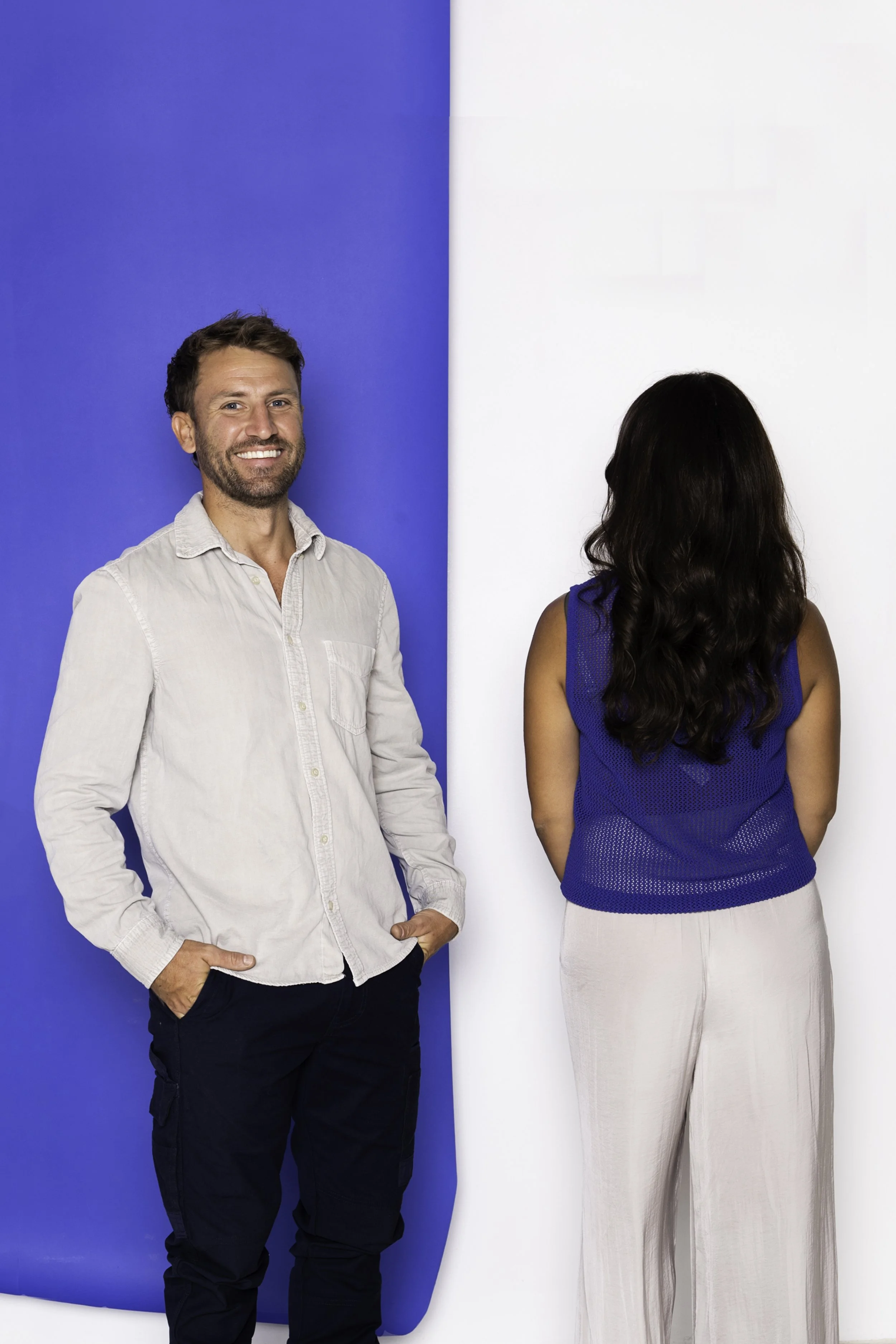 A man with a beard and a woman with long dark hair stand in front of a split-colored background, blue on the left and white on the right, with the woman facing away from the camera and the man smiling and looking at the camera.