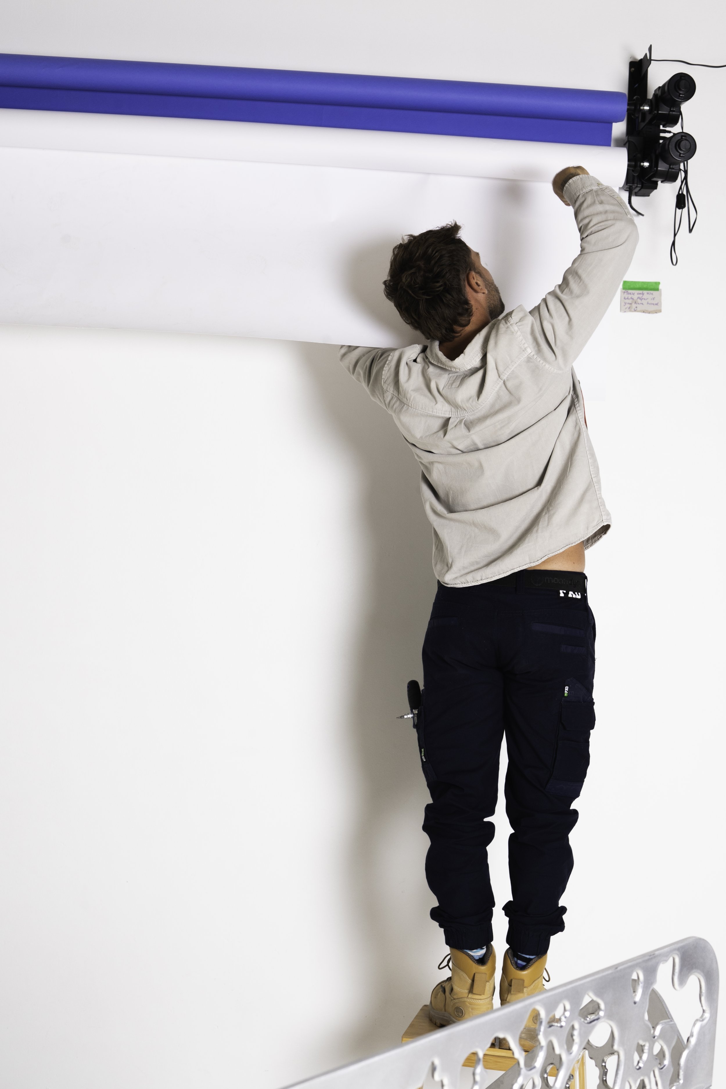 A man standing on a wooden platform, reaching up towards a wall with photo equipment and blue and white backdrop rolls mounted above him.