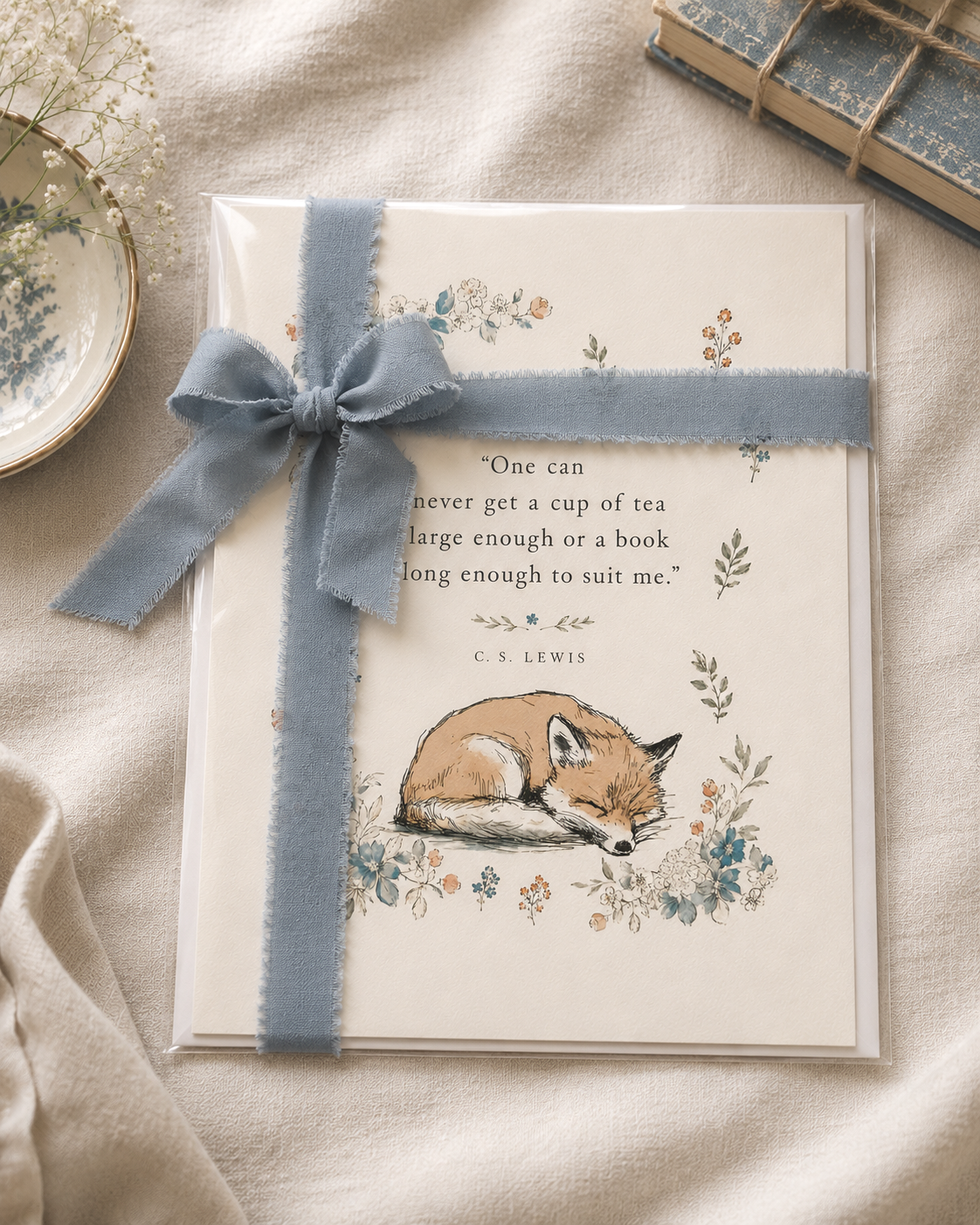 An art print with a quote by C.S. Lewis, an illustration of a sleeping fox, surrounded by floral designs, wrapped with a blue ribbon and bow, alongside a dish with flowers and a stack of tied books on a beige fabric background.